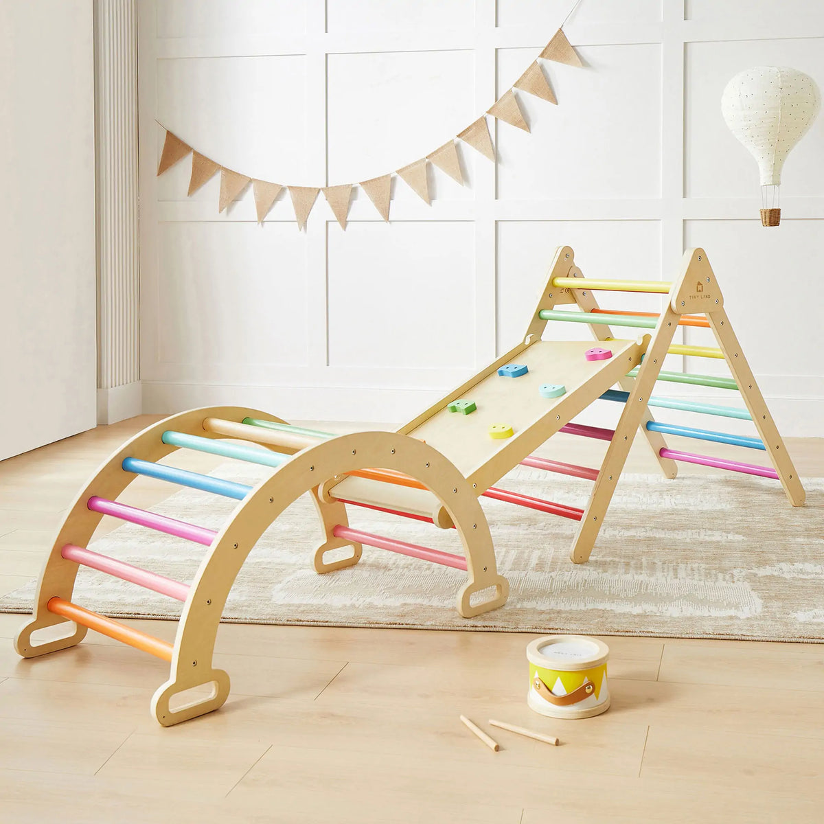 The Tiny Land® 7-in-1 Rainbow Climbing Set by Tiny Land includes a Pikler Triangle, wooden arch ladder, and slanted climbing board with colorful grips. A toy drum and stick also sit on the light wood floor in this bright room.|rainbow color