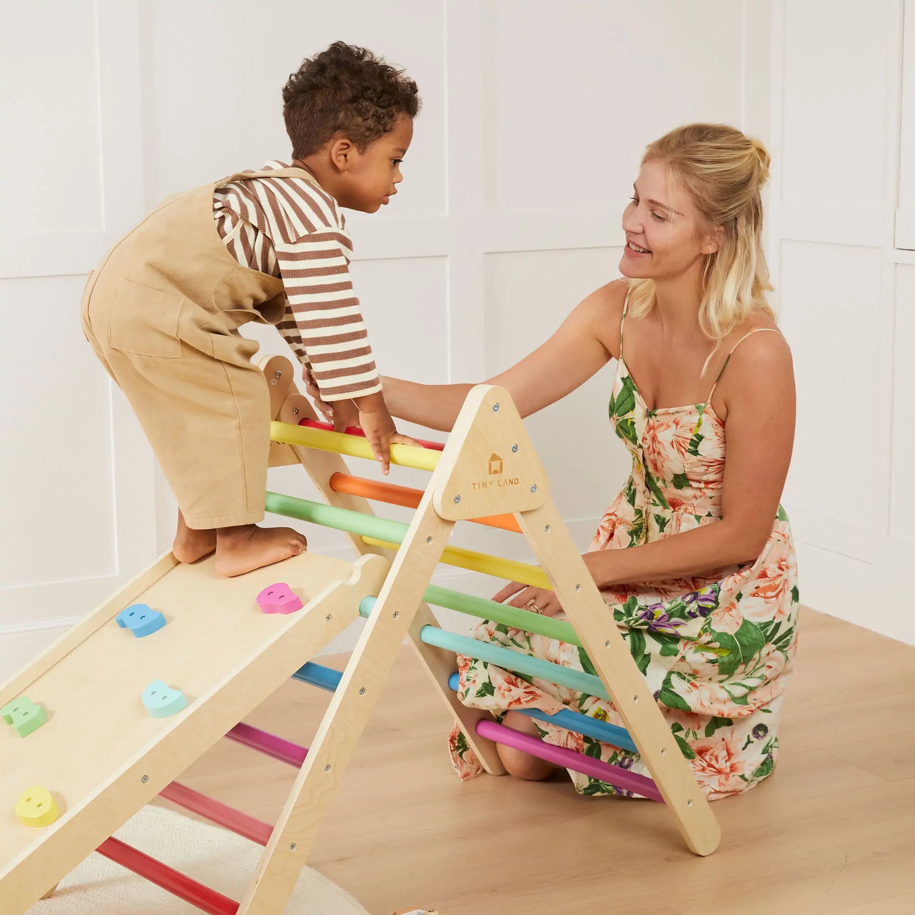A young child climbs the Tiny Land® 7-in-1 Rainbow Climbing Set as an adult woman in a floral dress kneels nearby, smiling and supporting the child in a bright, minimal room.