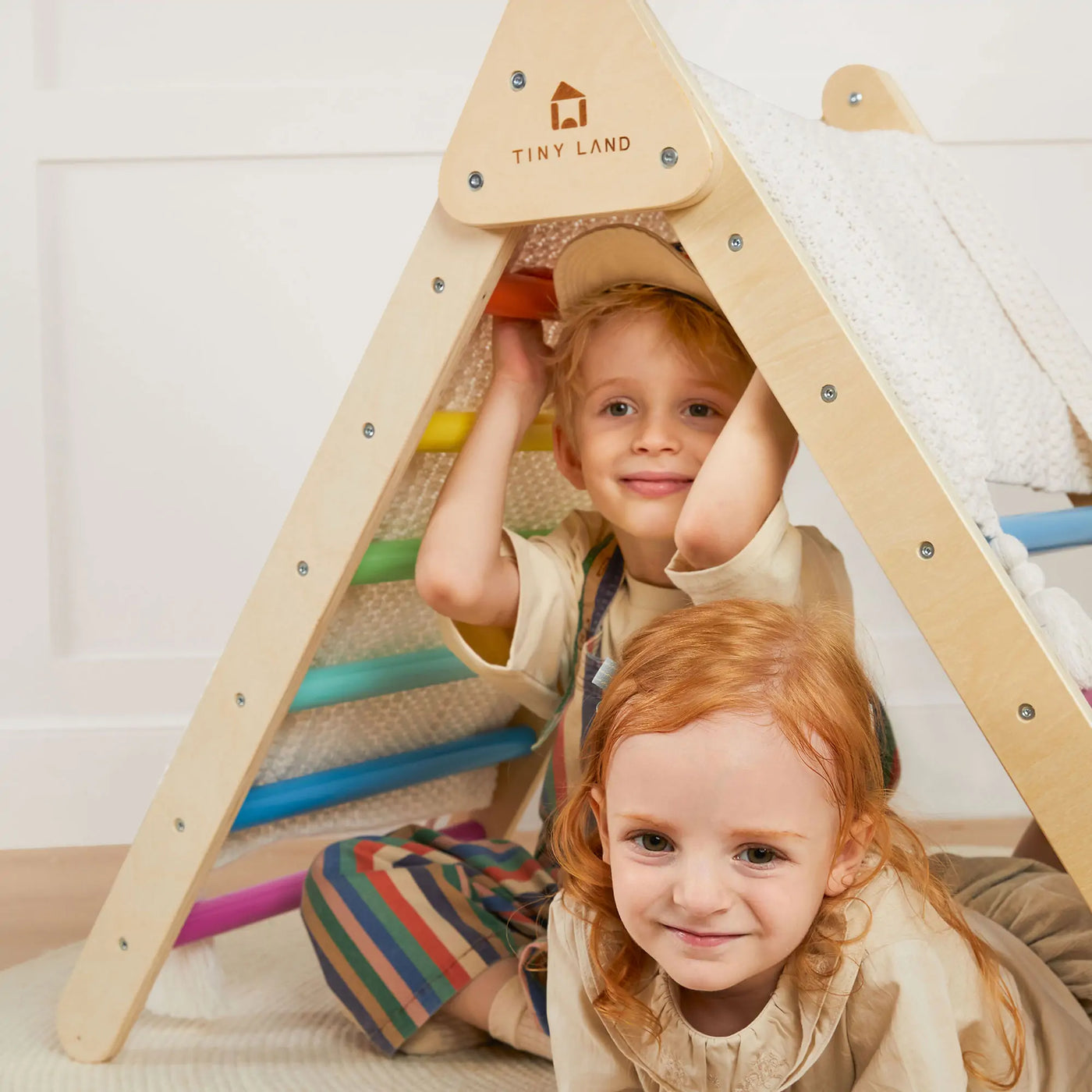 Two young children play under the Tiny Land Adventure Playground Gift Set, featuring a climbing set with pad, balance beam, and stepping stones. One sits inside the triangle smiling while the other crouches nearby, both happy and engaged in imaginative play.