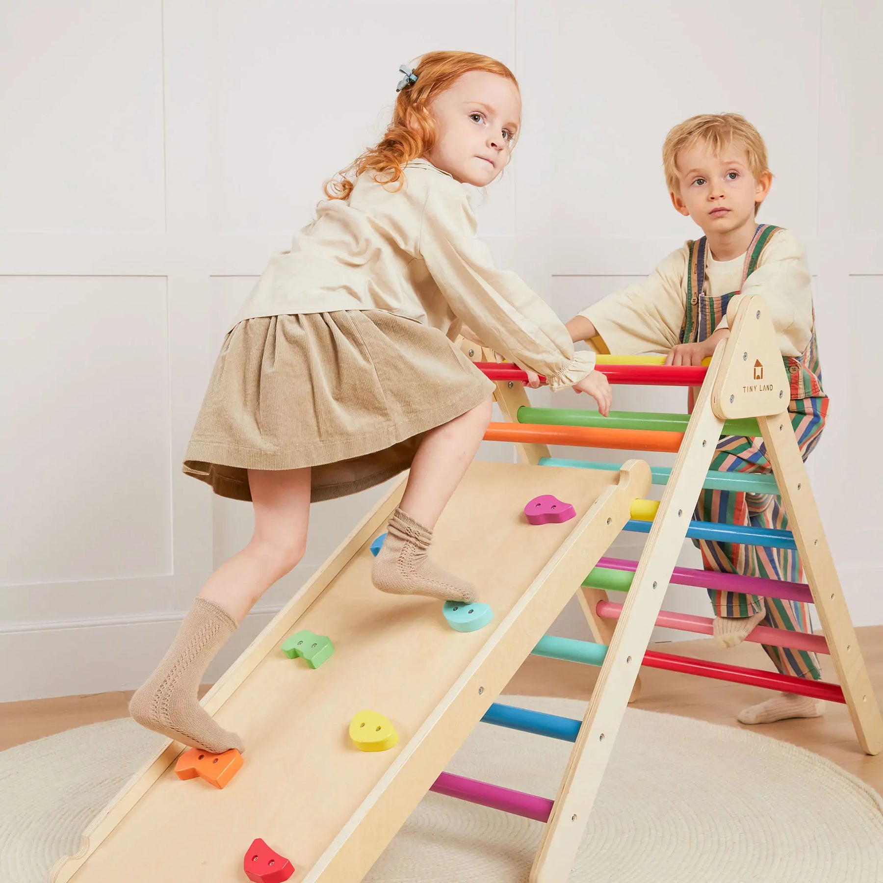 Two young kids play on the Tiny Land® 7-in-1 Rainbow Climbing Set with Play Pad. One climbs the ramp while the other stands at the top, both focused and engaged, as the padded play pad by Tiny Land adds comfort to their indoor adventure.