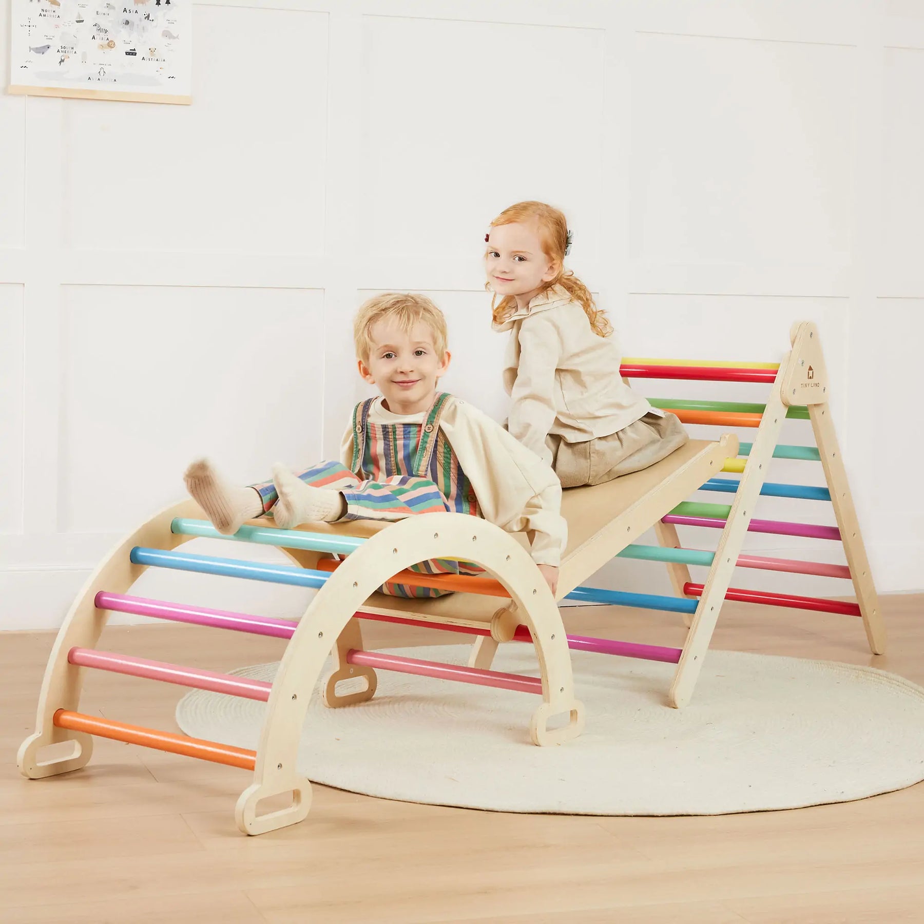 Two children play and smile on the Tiny Land® 7-in-1 Rainbow Climbing Set with Play Pad, featuring a Pikler triangle, arch, ramp, and colorful rungs by Tiny Land in a bright room with a padded play cushion beneath them.