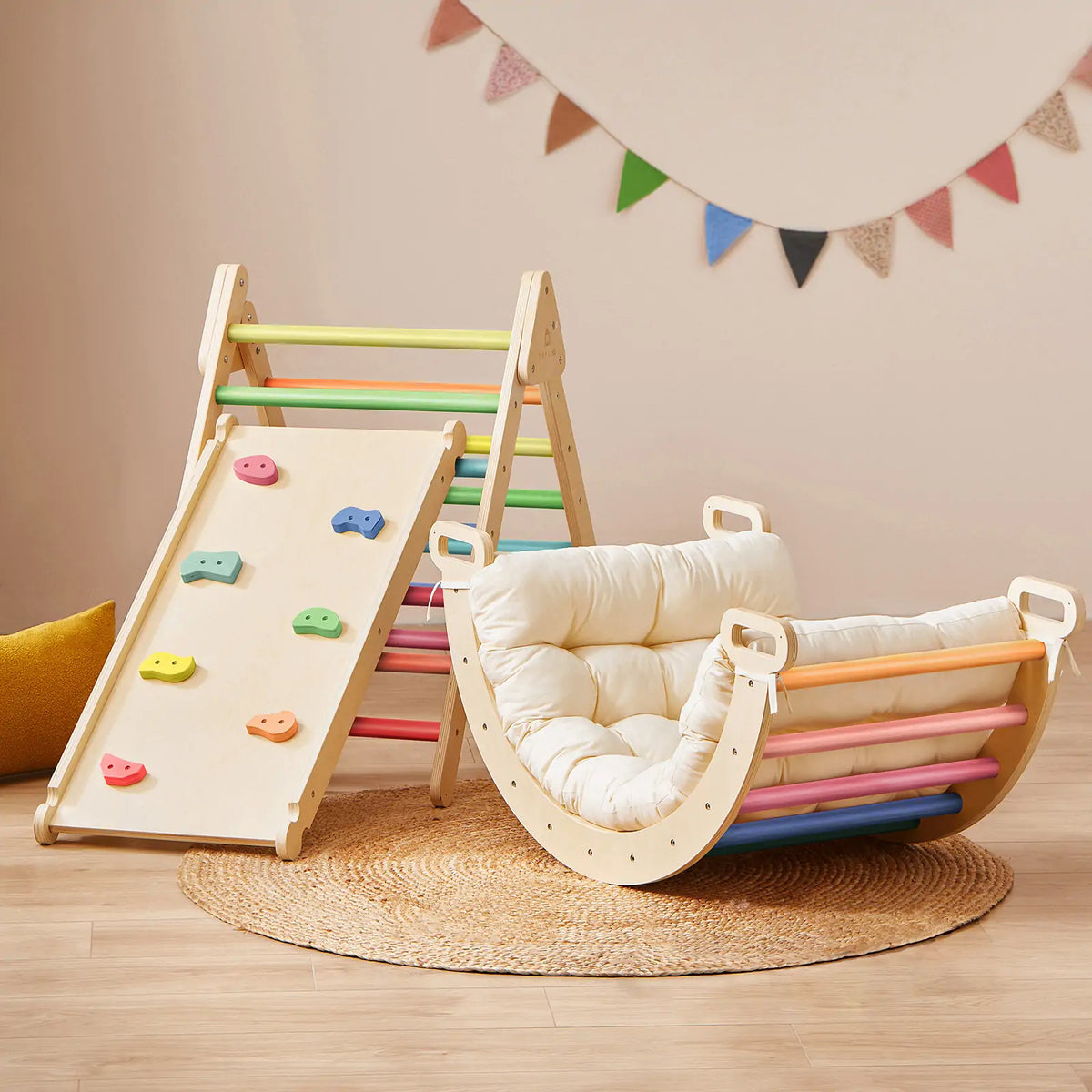 The Tiny Land Adventure Playground Gift Set, featuring a wooden Montessori climbing set with ramp and colorful rungs, sits beside a padded rocker cushion on a round woven rug in a cozy playroom with bunting on the wall.