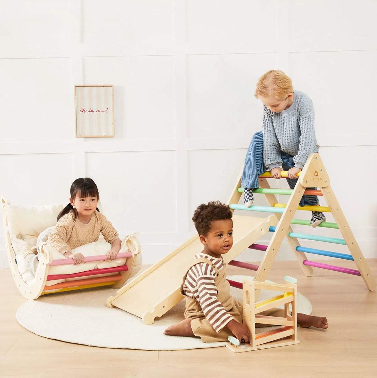 Three young children play indoors with the Tiny Land Adventure Playground Gift Set, including a climbing set with pad, balance beam, and stepping stones. One climbs the ramp while others explore cozy play cushions on a bright room's round rug.