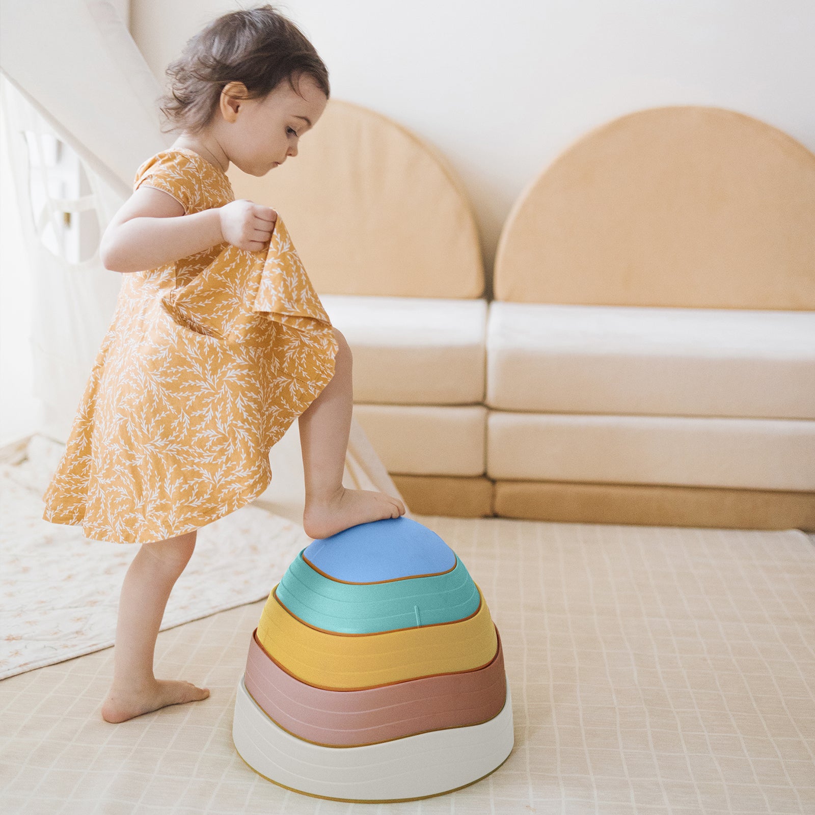 A young child in a yellow dress plays on the Tiny Land® Stepping Stones - Beach Vacation set, building motor skills in a cozy room with beige furniture and a patterned mat by creating a fun obstacle course.
