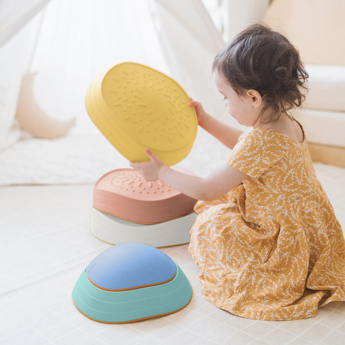 A young child in a yellow patterned dress sits on the floor of a cozy room, stacking colorful Tiny Land® Stepping Stones - Beach Vacation, building motor skills through fun obstacle play with these textured stones by Tiny Land.|Beach Vacation
