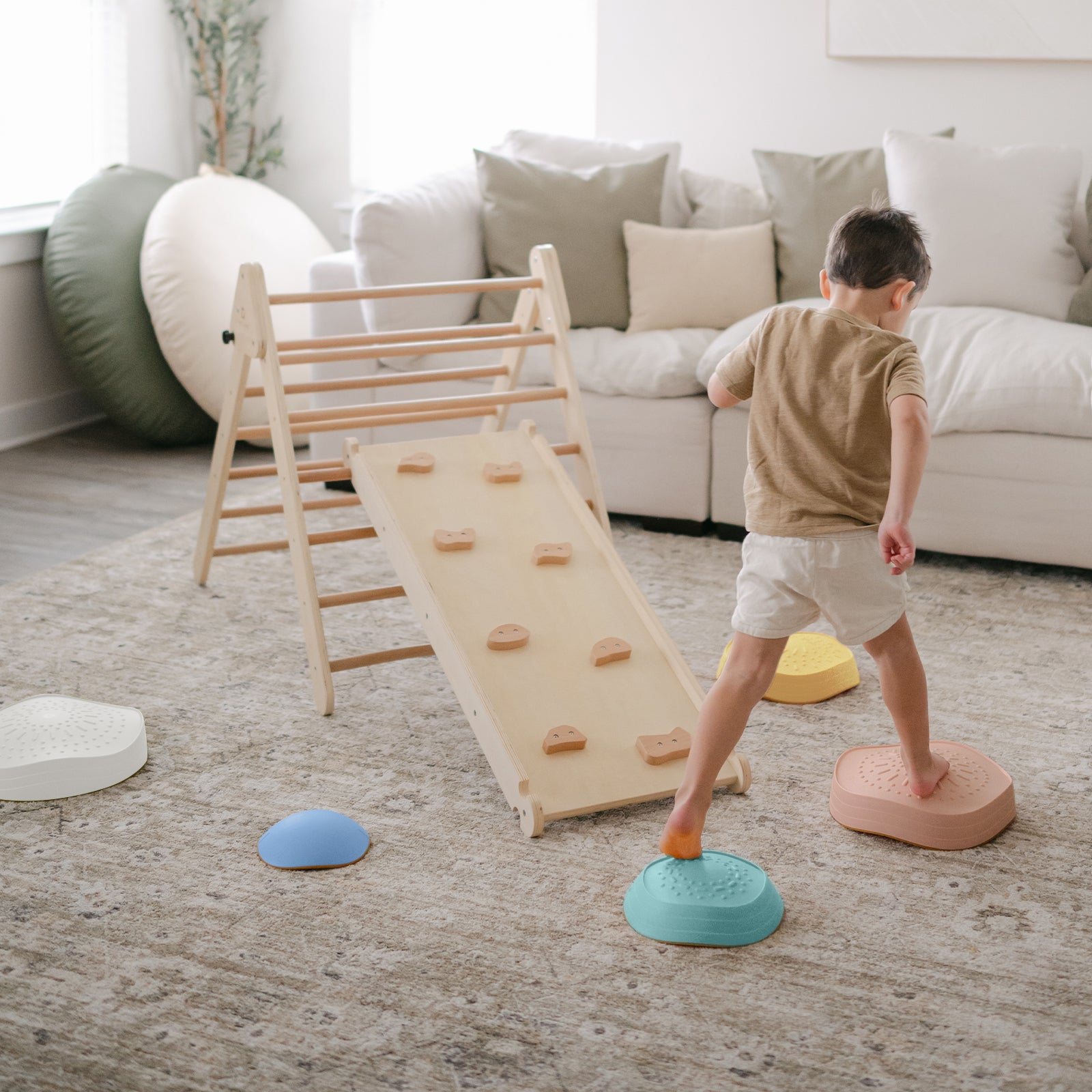 A young child steps on Tiny Land® Stepping Stones - Beach Vacation by Tiny Land, creating an indoor obstacle course toward a wooden climber with ramp in a cozy, well-lit living room to build motor skills.