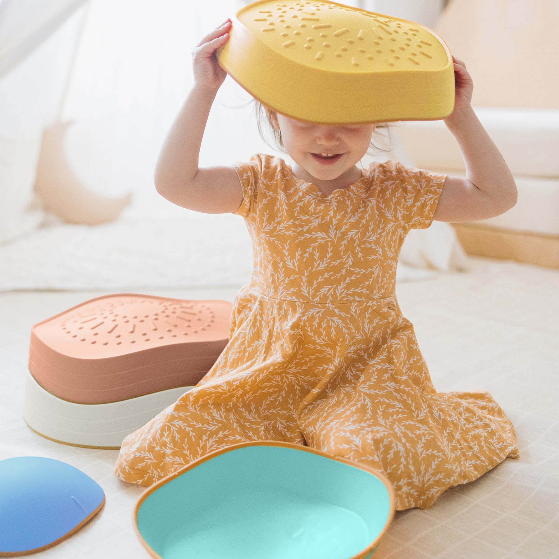 A young child in a yellow patterned dress sits on the floor with an upside-down yellow colander on their head. Tiny Land® Stepping Stones - Beach Vacation by Tiny Land are scattered around, perfect for obstacle course fun and motor skill development.