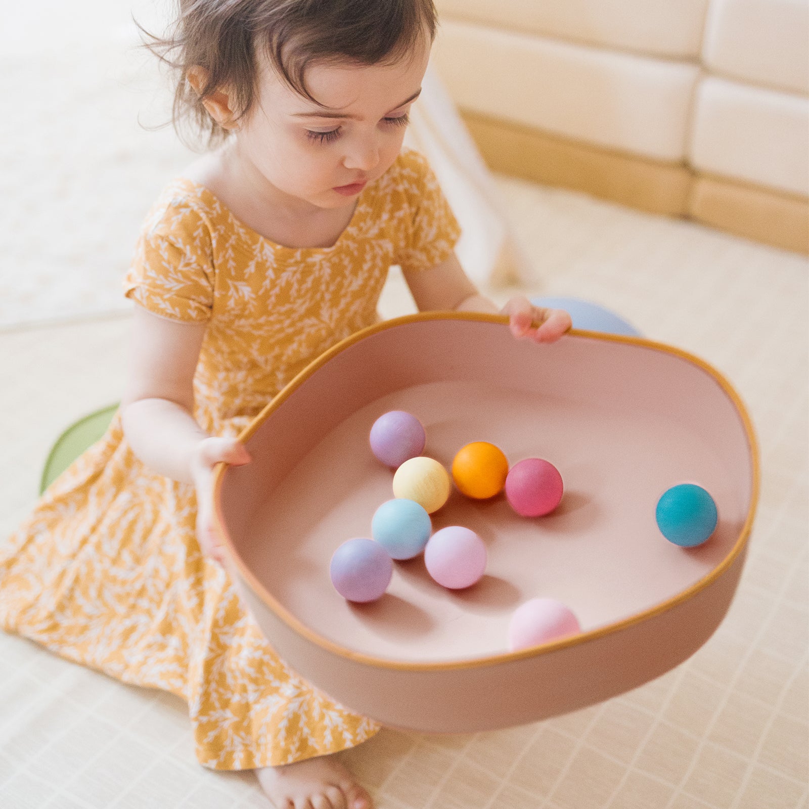 A young child in a yellow dress sits indoors on a light floor, holding a large pink bowl of colorful balls—perfect for developing motor skills after playing on the Tiny Land® Stepping Stones - Beach Vacation by Tiny Land.