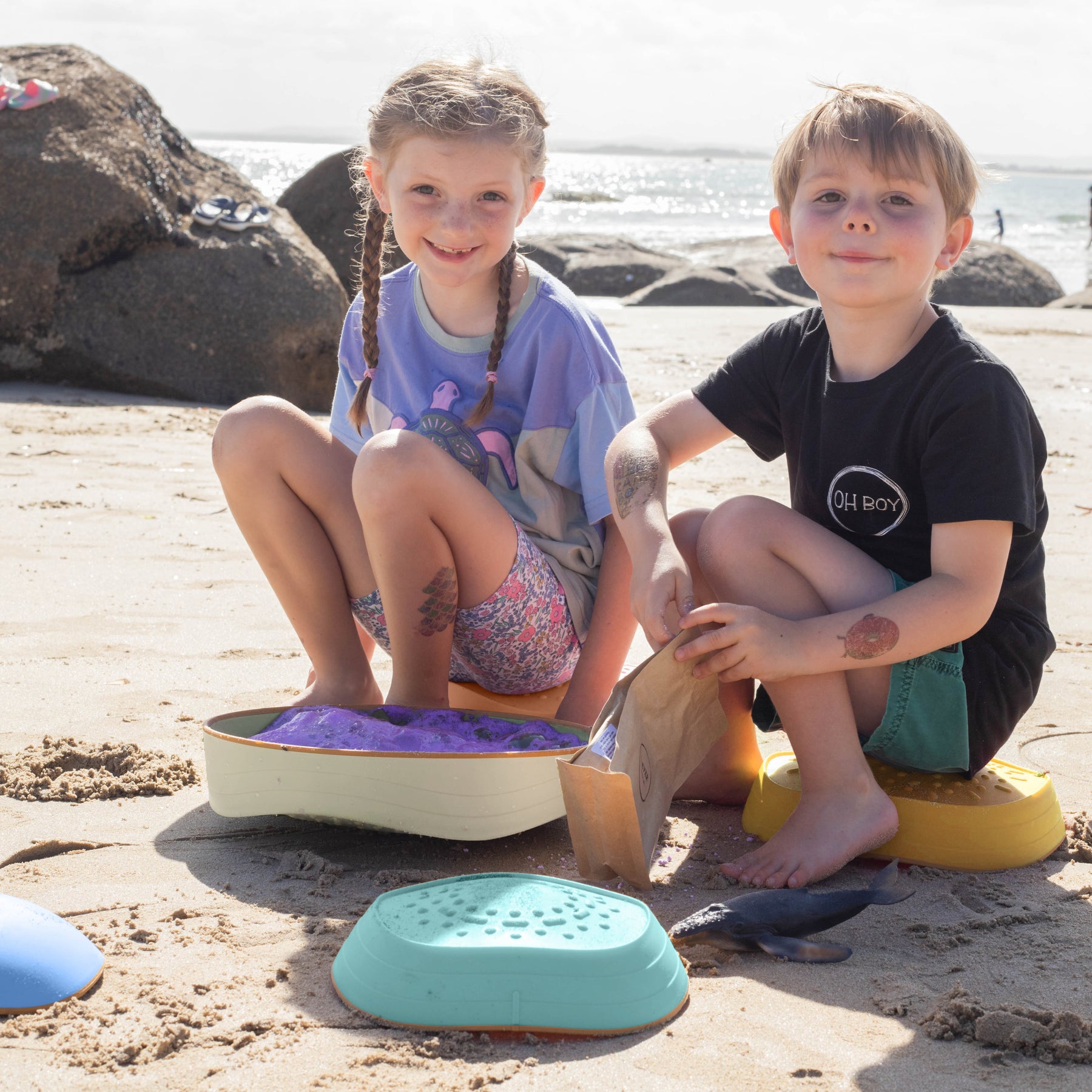 Two young children smile and play on the beach with sand toys and Tiny Land® Stepping Stones - Beach Vacation, building motor skills as waves sparkle behind them under a sunny sky.