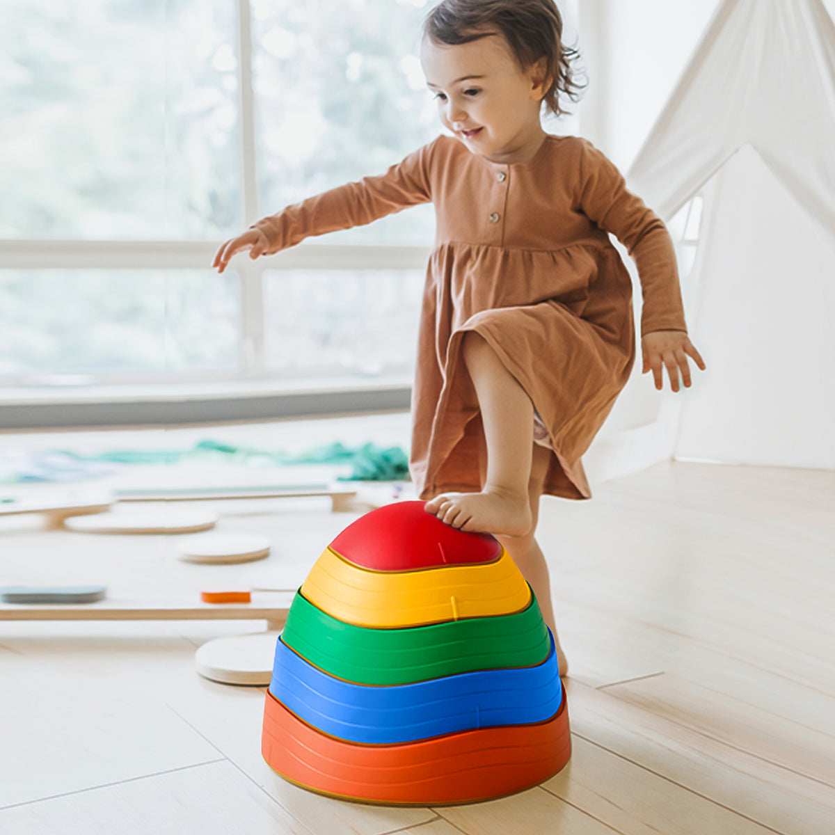 A young child in a brown dress balances barefoot on Tiny Land’s Adventure Playground Gift Set—featuring a climbing set with pad, balance beam, and stepping stones—in a bright, modern playroom with wooden floors and large windows.|Rainbow Color