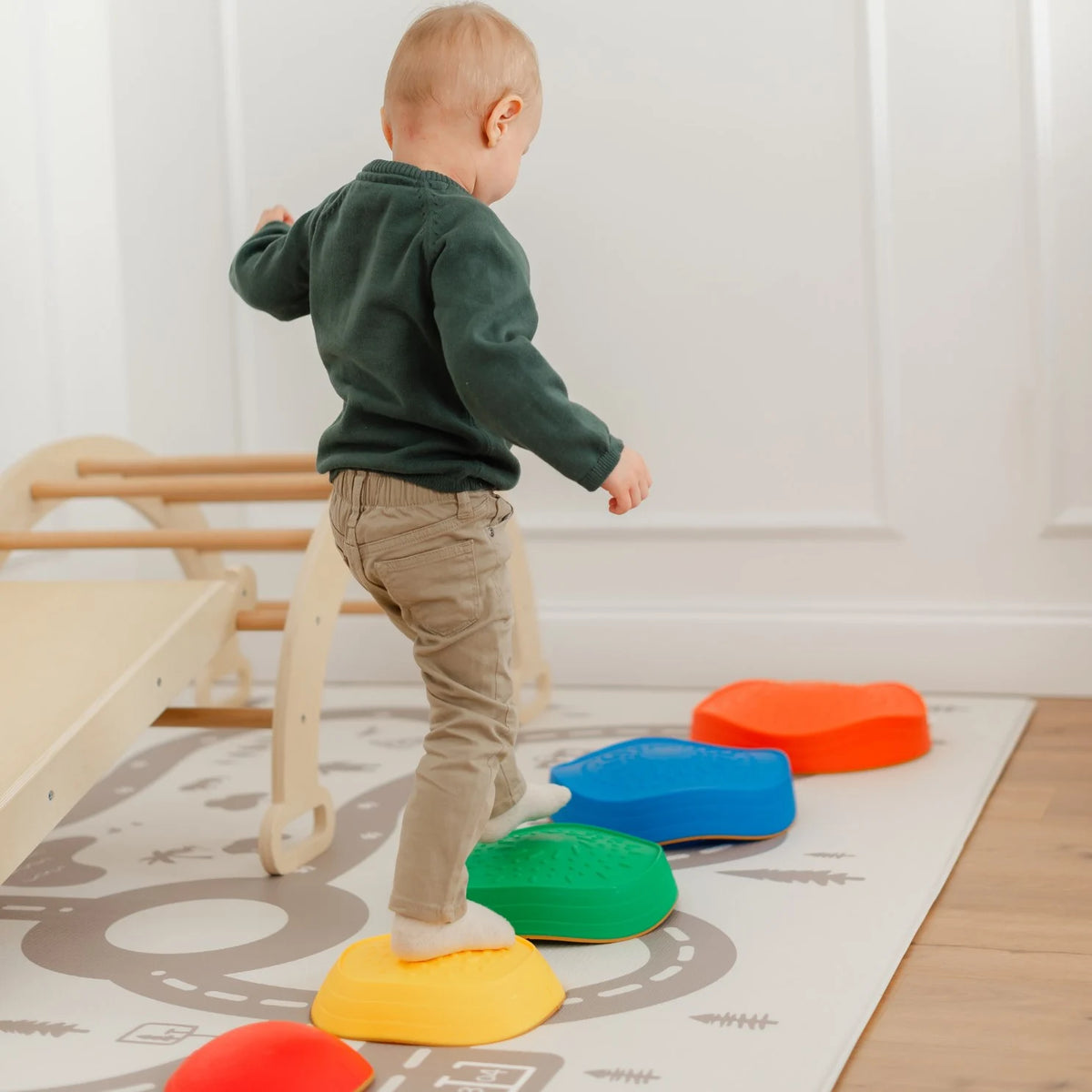 A young child in a green sweater and beige pants balances on Tiny Land's Adventure Playground Gift Set stepping stones, arranged on a play mat beside the climbing set in a bright indoor room.