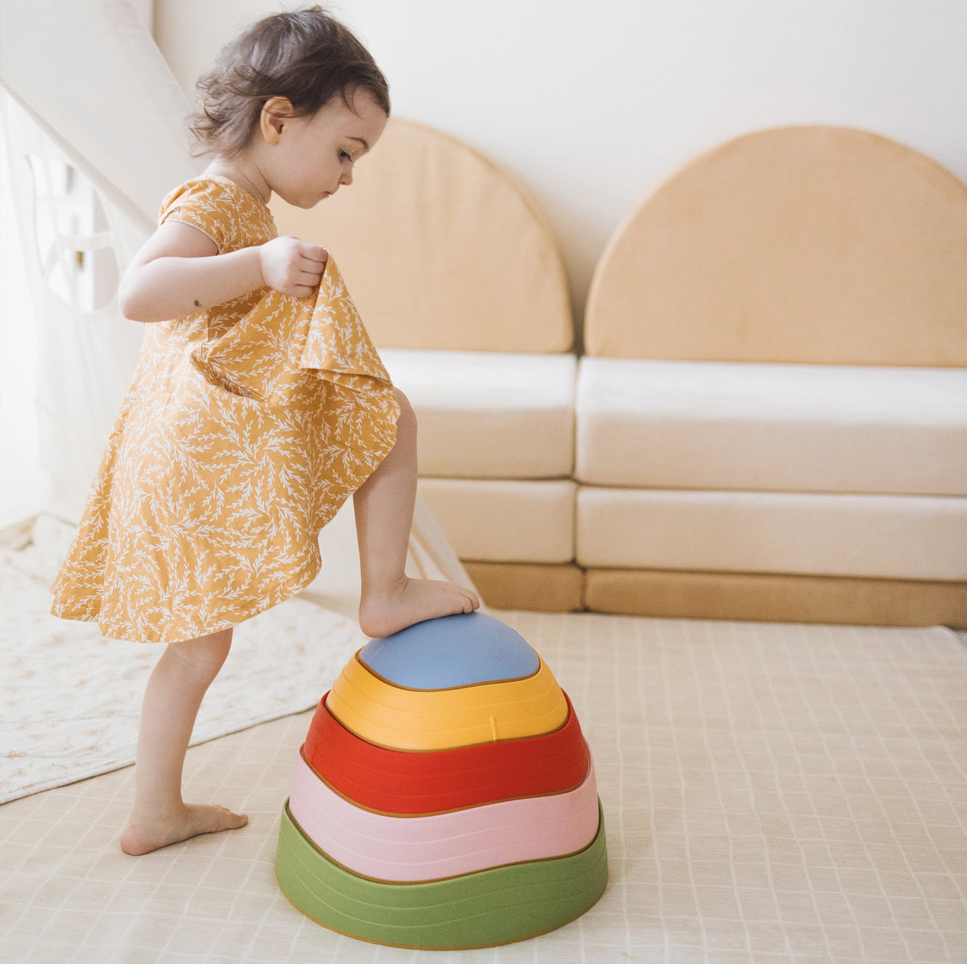 A young child in a yellow dress steps onto Tiny Land® Stepping Stones - Vintage Color, building motor skills in a bright playroom with a beige couch and soft mat.