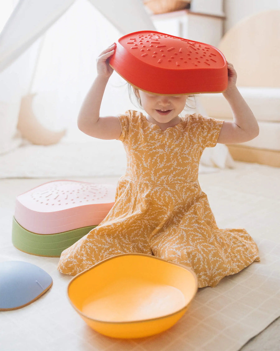 A young child in a yellow dress sits on the floor, smiling as they play with a red bowl beside Tiny Land® Stepping Stones - Vintage Color. The colorful stepping stones by Tiny Land are scattered across the light-colored floor.|Vintage Color