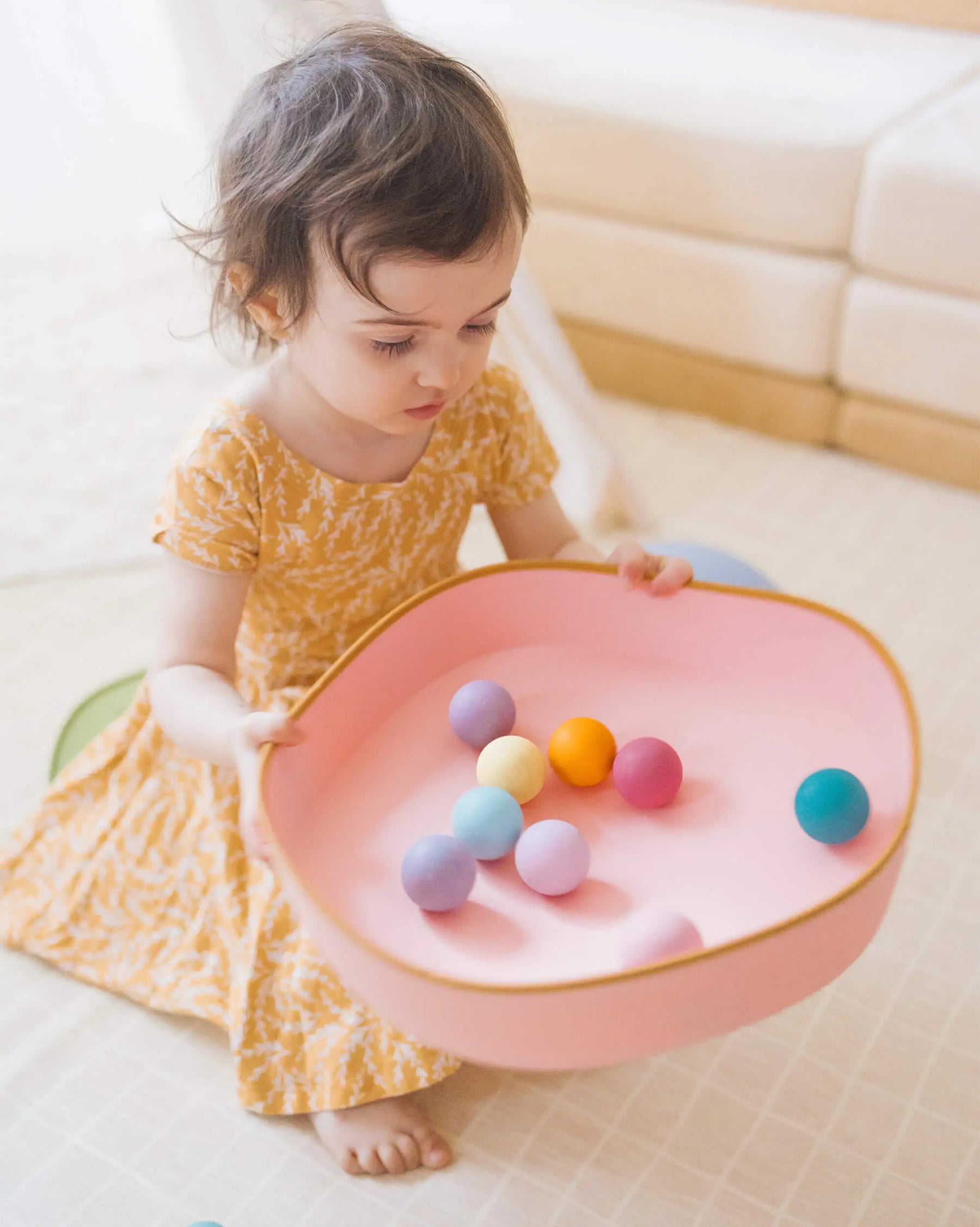 A young child in a yellow dress kneels on a light floor indoors, building motor skills as she plays with colorful balls near the Tiny Land® Stepping Stones - Vintage Color by Tiny Land.