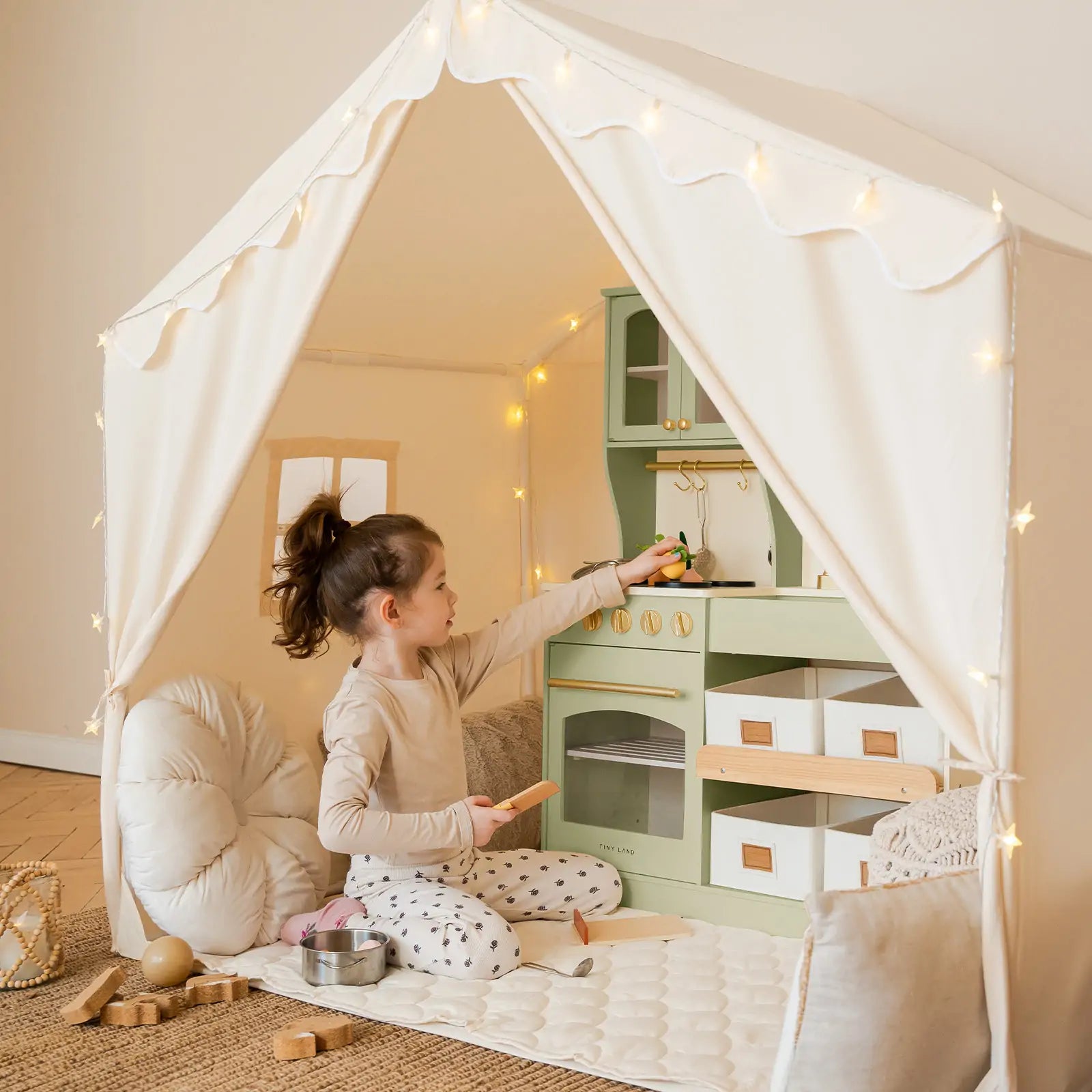 A young girl sits inside the Tiny Land® Kids Tent – Large Cozy Tent with Star Lights by Tiny Land, playing with a toy kitchen set and surrounded by soft cushions and wooden toys on a quilted play mat.