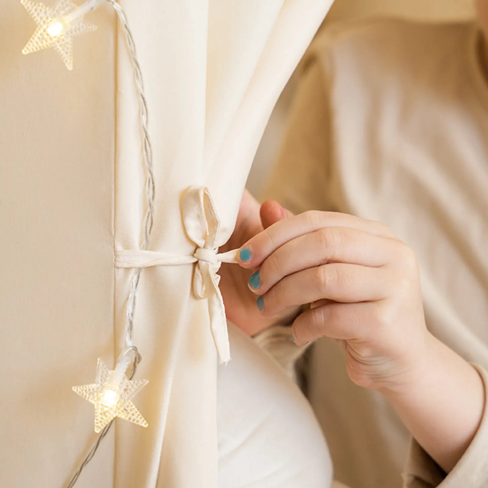 A person with blue nail polish ties a white ribbon around the Tiny Land® Kids Tent – Large Cozy Tent with Star Lights by Tiny Land; star-shaped lights and a quilted play mat nearby create a cozy, softly lit atmosphere.