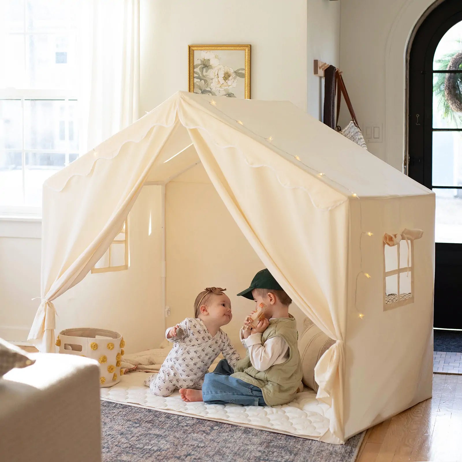 Two children play in a sunlit living room inside the Tiny Land® Kids Tent – Large Cozy Tent with Star Lights. One wears pajamas, the other a cap and vest. A basket and quilted mat add comfort to this inviting scene by Tiny Land.
