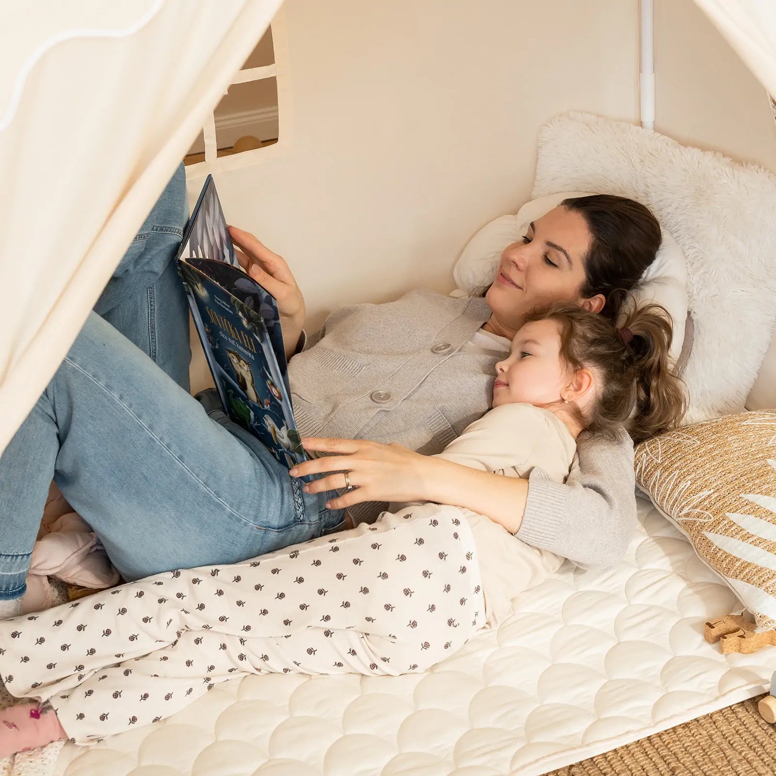 A woman and a young girl relax inside the Tiny Land® Kids Tent – Large Cozy Tent with Star Lights, reading together. The cozy hideaway from Tiny Land offers comfort and warmth, perfect for special bonding moments.