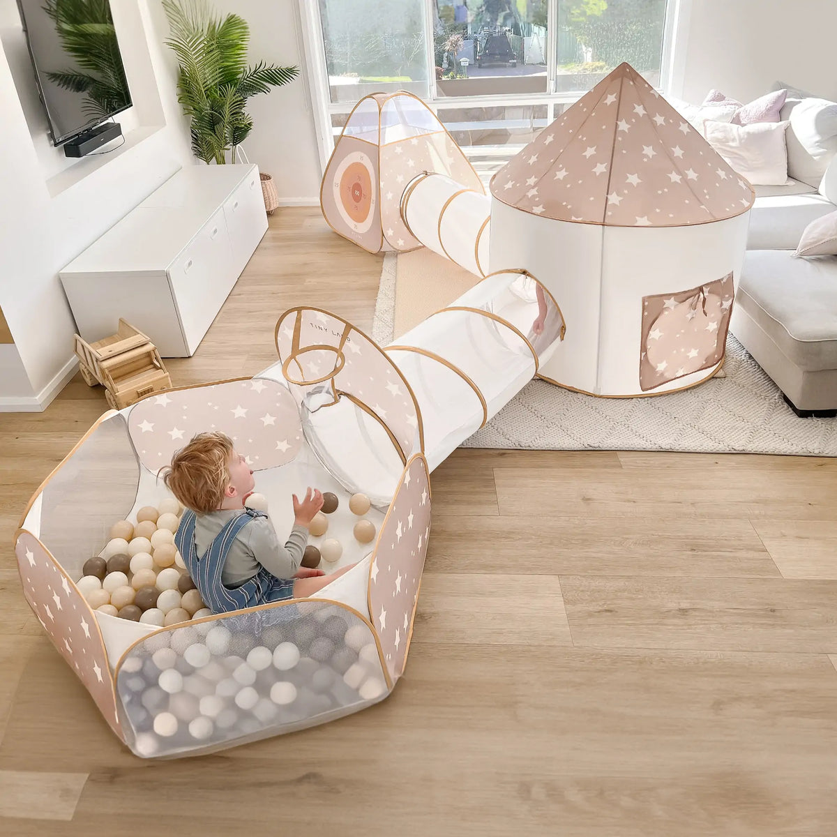 A toddler enjoys the Tiny Land® Mystery Surprise Box—a beige and white indoor play tent set with a ball pit, tunnel, and star patterns—by Tiny Land, set up in a bright living room with wooden floors and a large window.