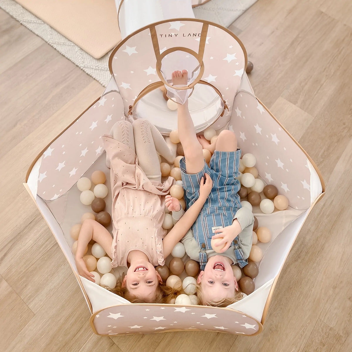 Two young children smile and relax in the Tiny Land® Discovery 5-in-1 Play Tunnel & Ocean Ball Set, featuring a pastel playpen with star patterns and beige, white, and brown ocean balls by Tiny Land. Perfect for active, joyful play.