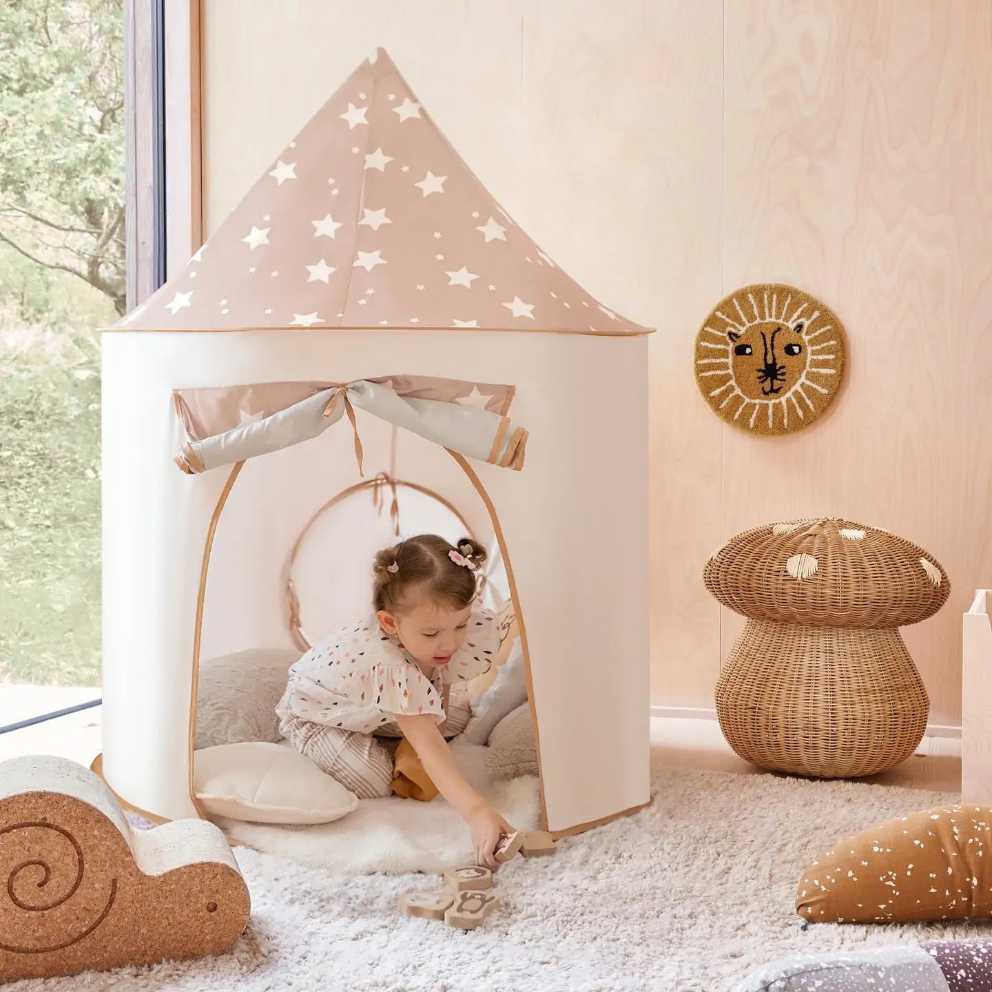 A young girl plays in a beige tent with a star-patterned roof. The Tiny Land® Discovery 5-in-1 Play Tunnel is nearby, inviting adventure. The room features a plush rug, wicker lion decor, mushroom stool, and a window with greenery outside.