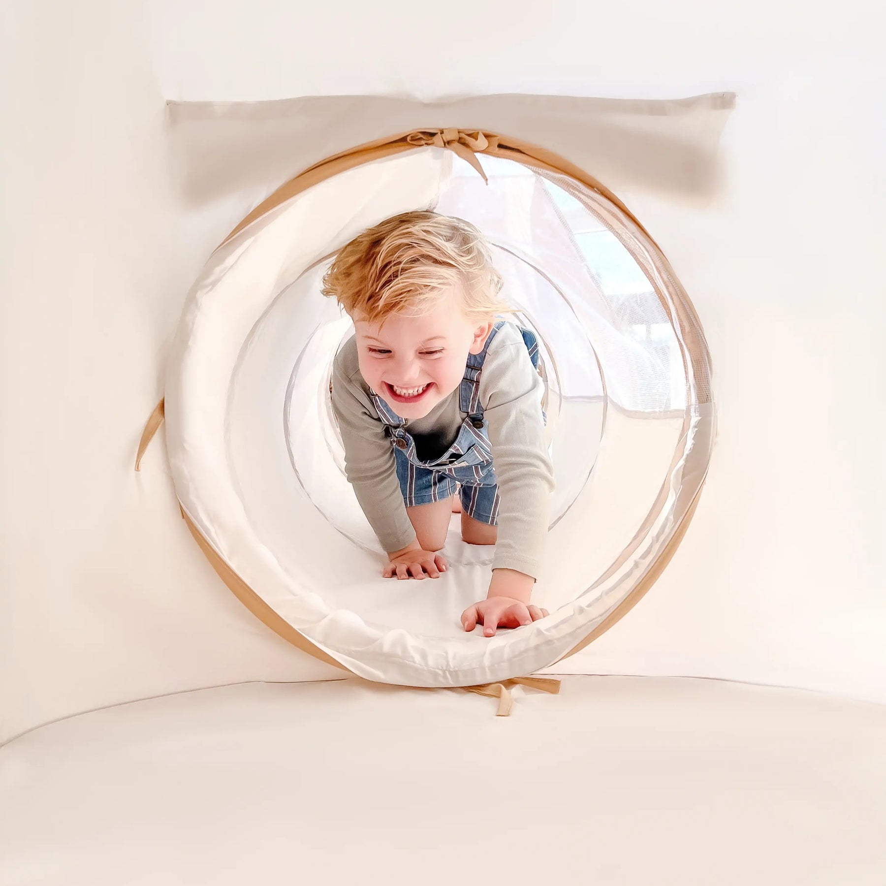A young child with blonde hair and a grey shirt smiles while crawling in the Tiny Land® Discovery 5-in-1 Play Tunnel (no balls) by Tiny Land, set in a bright, light-colored room.