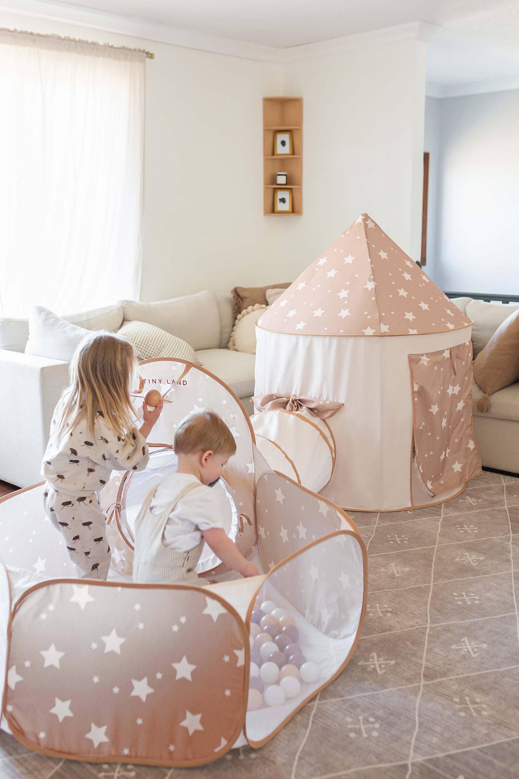 Two young children play in the Tiny Land® Compact Adventure 3-in-1 Play Tunnel, a star-patterned tent set up in a bright living room with a beige couch and sheer curtains.