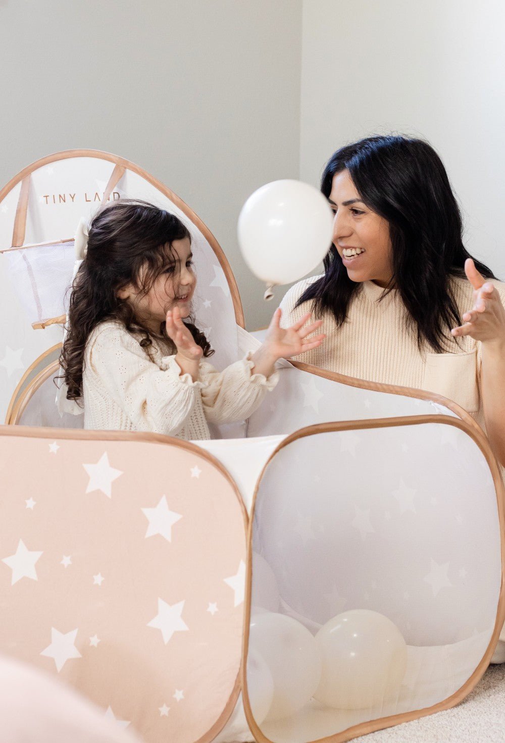 A woman and girl with dark hair smile and play with a white balloon inside the Tiny Land® Compact Adventure 3-in-1 Play Tunnel, surrounded by scattered balloons that turn the tent into their own playful ball pit.