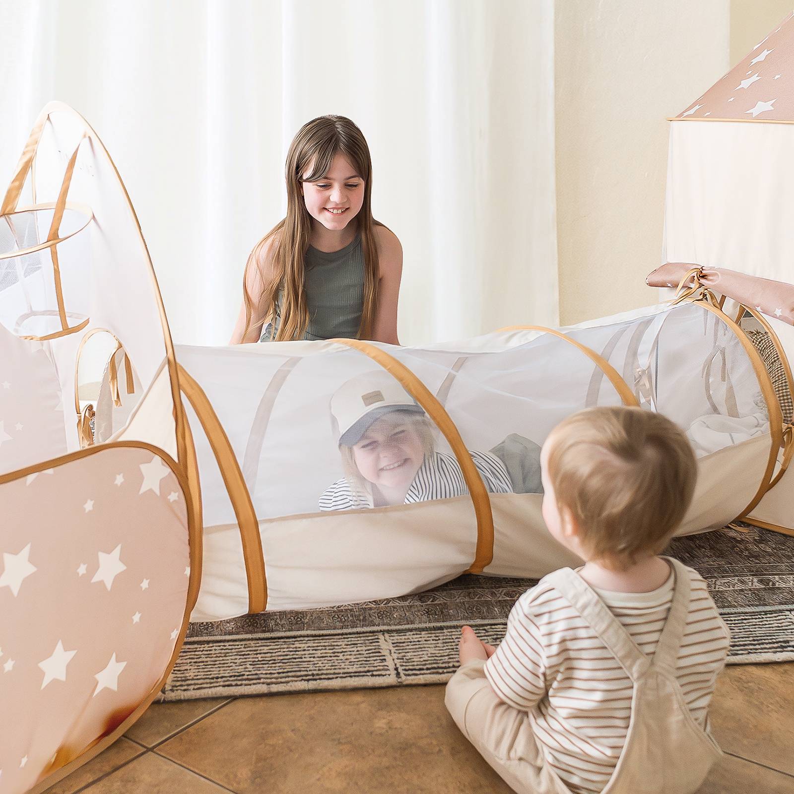 A young girl smiles at two kids playing with the Tiny Land® Compact Adventure 3-in-1 Play Tunnel; one is inside, the other sits at the entrance, transforming their space into an imaginative tent.
