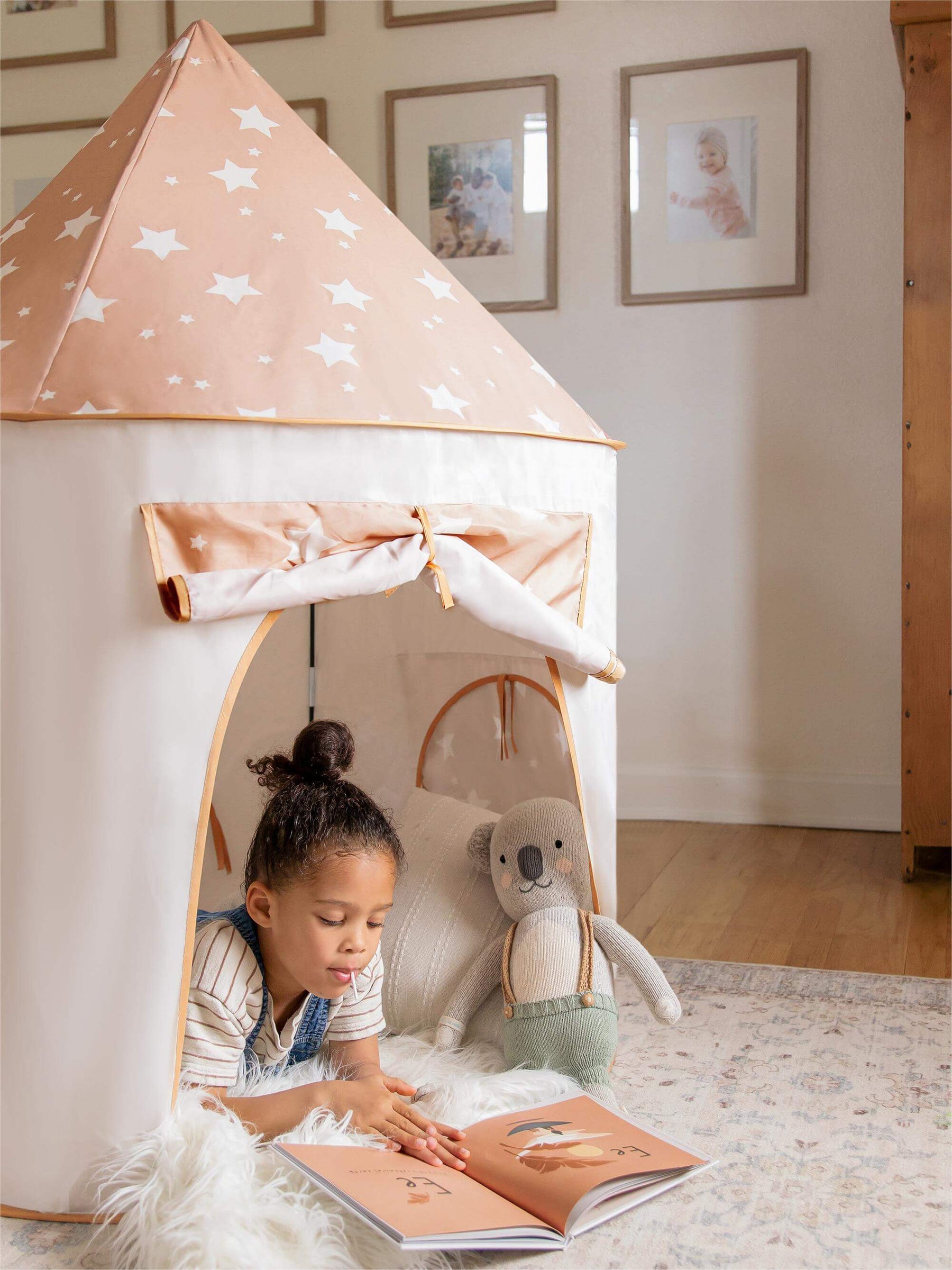 A young girl reads inside the Tiny Land® Adventure 3-in-1 Play Tunnel & Ocean Ball Set by Tiny Land, with a starry play tent, a stuffed animal on a cozy rug, and framed photos warming the room.