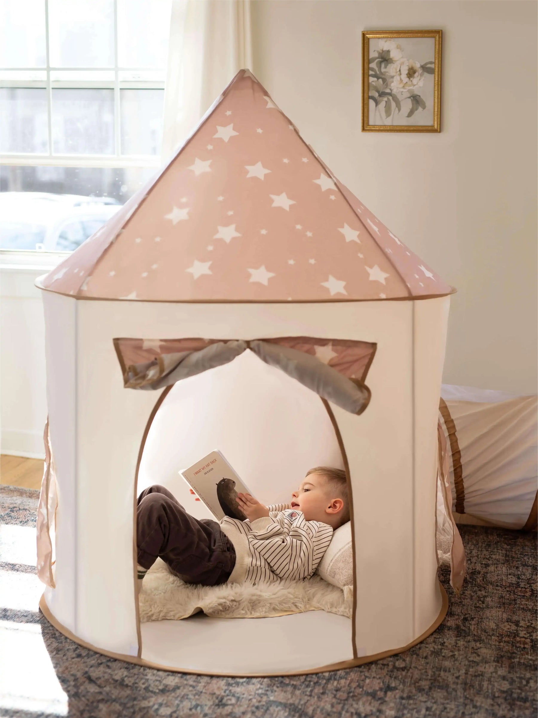 A young child relaxes inside the Tiny Land® Compact Adventure 3-in-1 Play Tunnel, reading a book. Soft light filters in, highlighting the playful space by Tiny Land, with a cozy atmosphere and a framed picture on the wall.