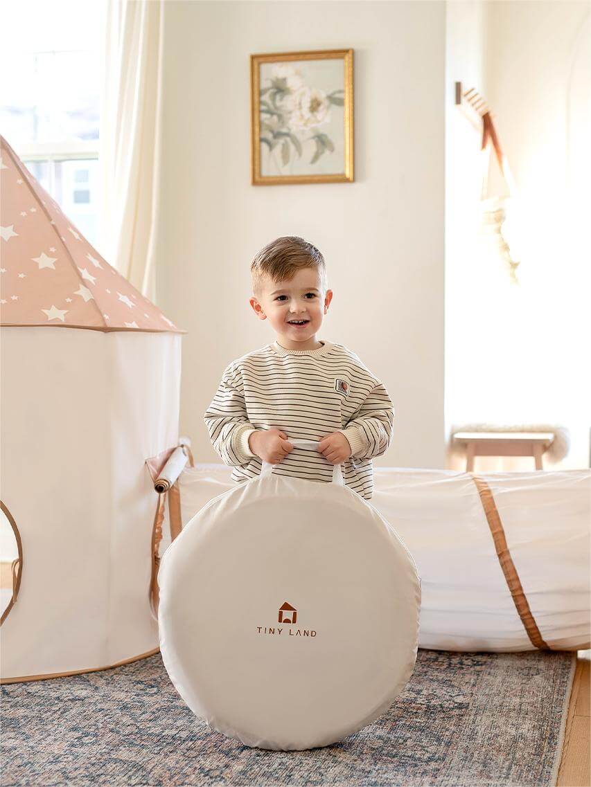 A young child smiles indoors, holding a Tiny Land storage bag. Behind them are the Tiny Land® Adventure 3-in-1 Play Tunnel & Ocean Ball Set, featuring a star-patterned tent, cozy ball pit, and soft play area in a bright room.