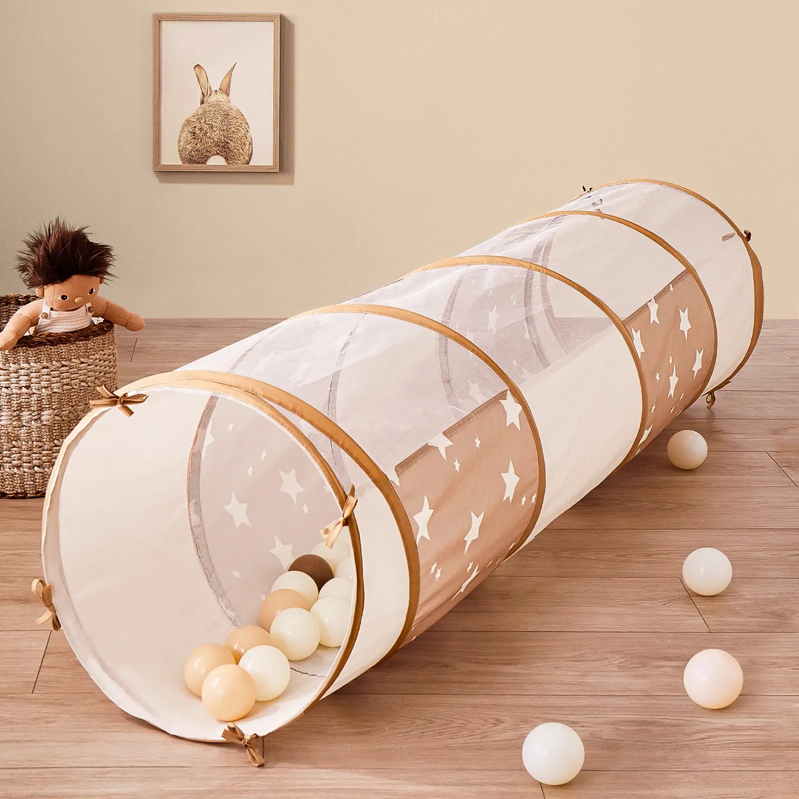 The Tiny Land® Adventure Play Tunnel, featuring mesh fabric and white stars, sits on a wooden floor with soft play balls. A plush doll in a woven basket and framed animal art decorate the beige wall in the background.