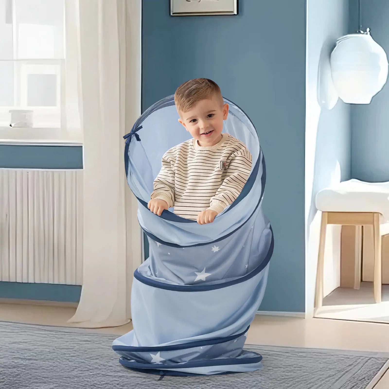 A smiling young boy stands inside the Tiny Land® Adventure Play Tunnel - Blue by Tiny Land, decorated with star patterns, in a bright room with light blue walls and a white radiator.