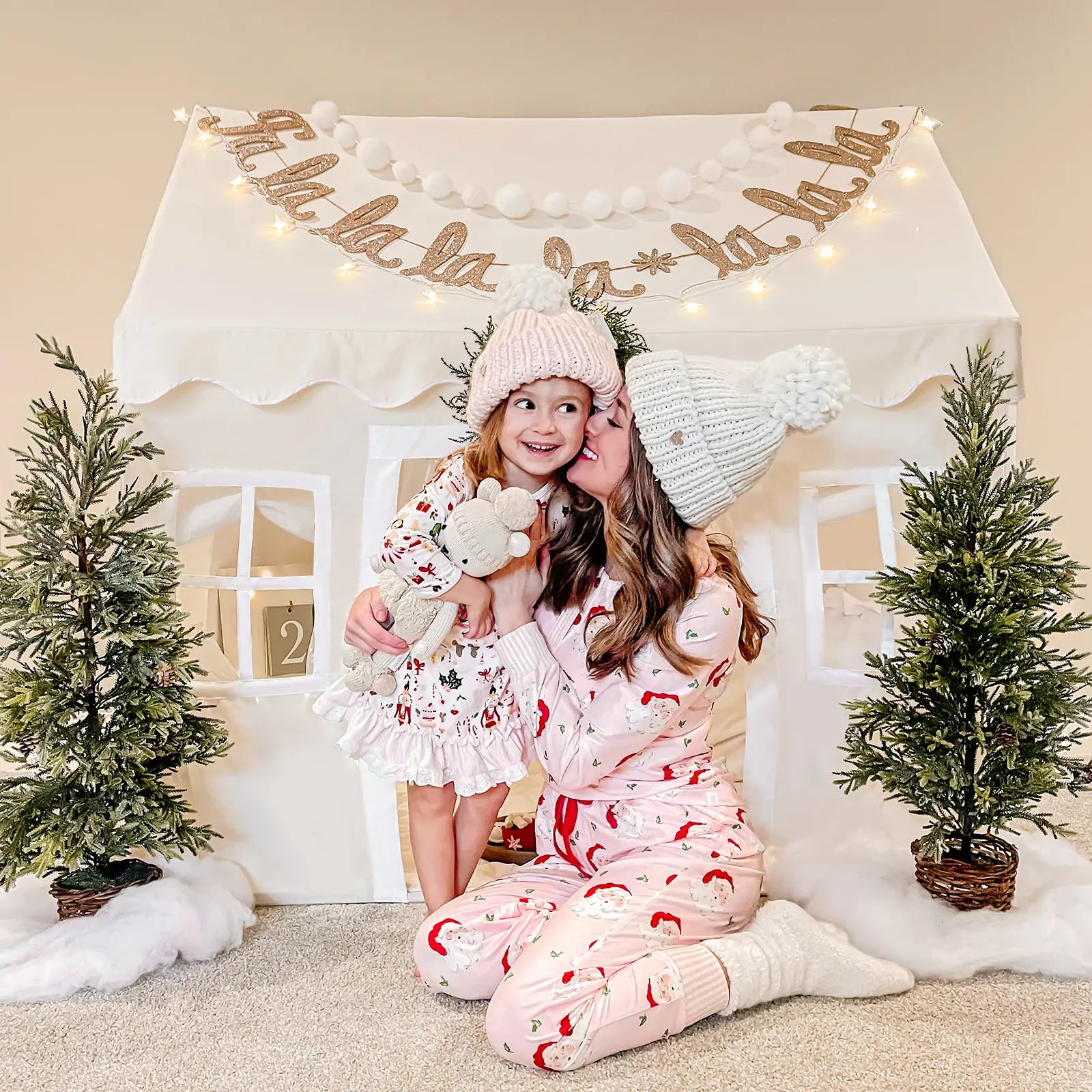 A woman in pink pajamas kisses a smiling young girl in a festive dress and knit hat as they sit together by the Tiny Land® Playhouse Tent – Natural Cream, surrounded by small Christmas trees and holiday décor.
