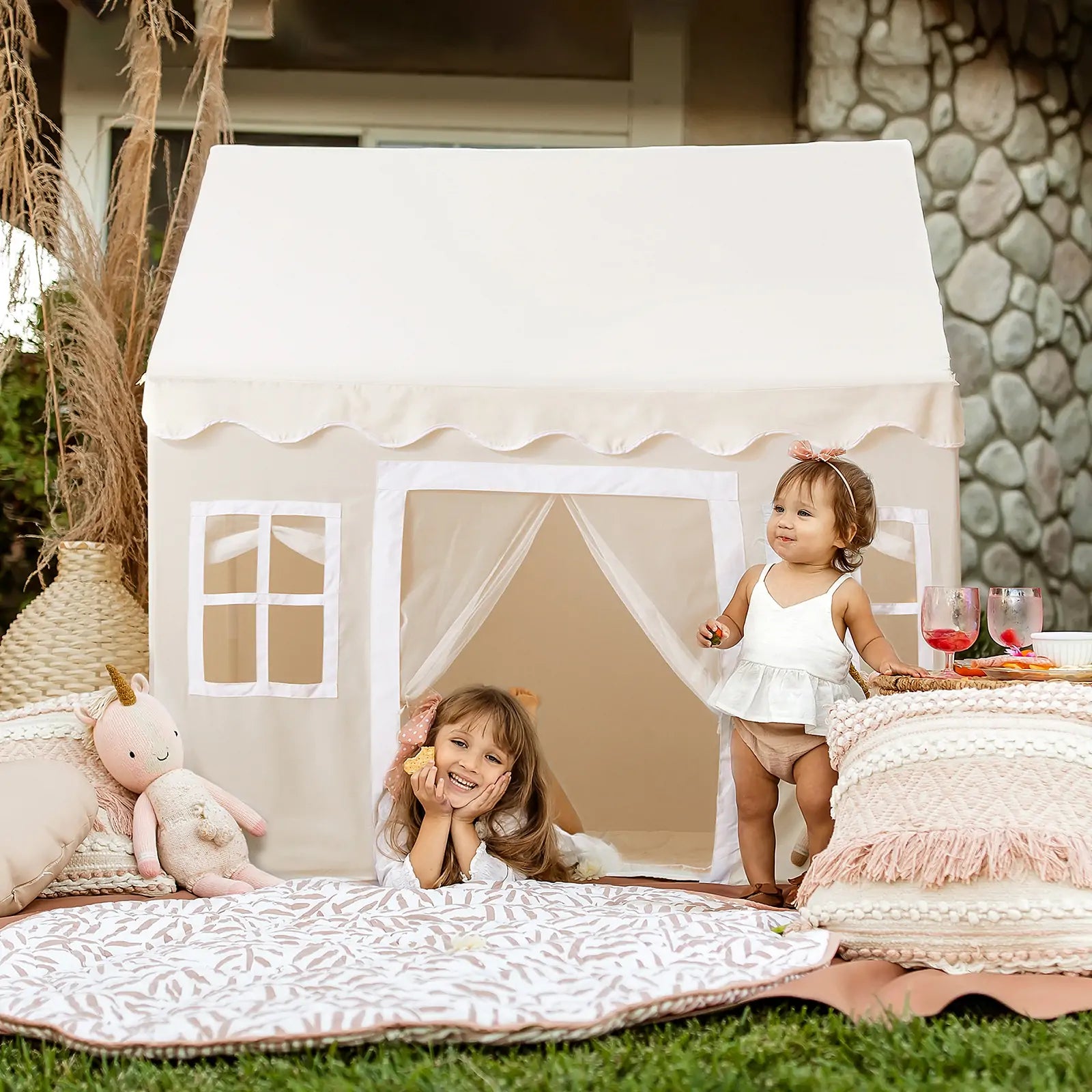 Two young children enjoy outdoor fun with the Tiny Land® Playhouse Tent – Natural Cream by Tiny Land. One smiles on a blanket while the other stands nearby, surrounded by plush cushions, a stuffed unicorn, and drinks.