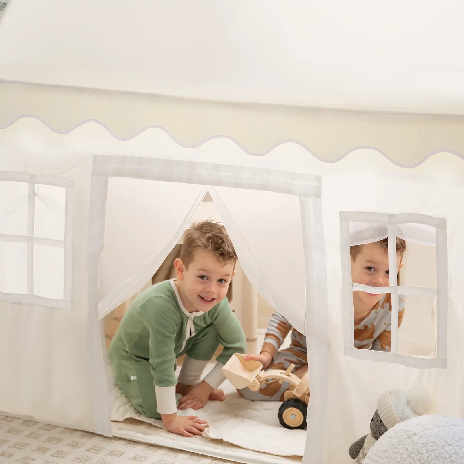 Two children play inside the Tiny Land® Playhouse Tent – a natural cream indoor tent by Tiny Land. One smiles at the entrance while the other peeks out, surrounded by soft toys and a wooden truck in this cozy scene.