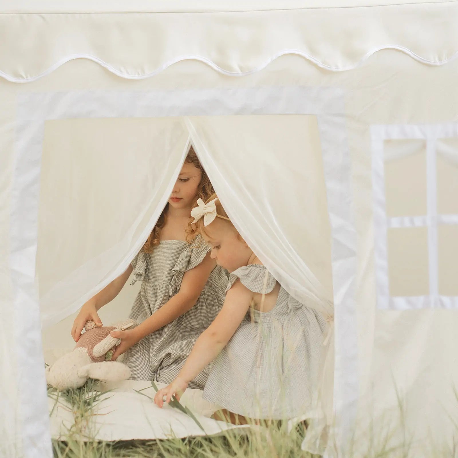 Two young children in matching dresses play inside the Tiny Land® Playhouse Tent – a natural cream indoor tent for kids by Tiny Land. One holds a stuffed bunny as sheer curtains and lush grass add to the cozy indoor-outdoor vibe.