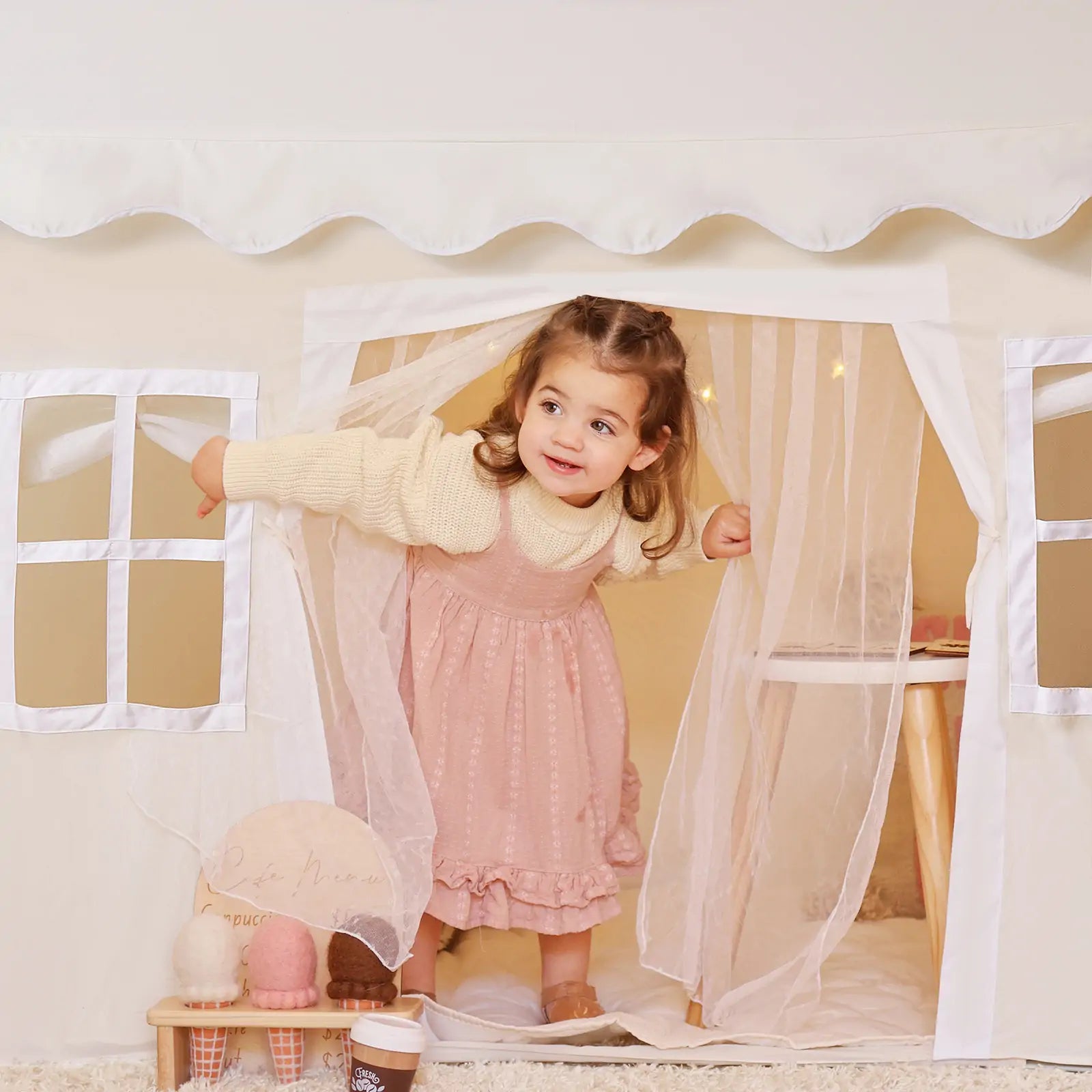A young girl smiles as she peeks out from the Tiny Land® Playhouse Tent – a natural cream indoor kids tent by Tiny Land, featuring sheer curtains, a small table, and décor for a cozy play space.