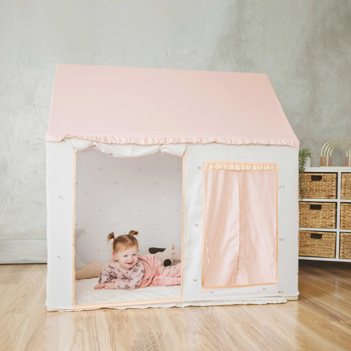 A young child in pink pajamas smiles while lying inside the Tiny Land® Sweet Citrus Kids Play Tent – a white and pink indoor playhouse by Tiny Land, set up on a wooden floor in a cozy, modern room.|pink