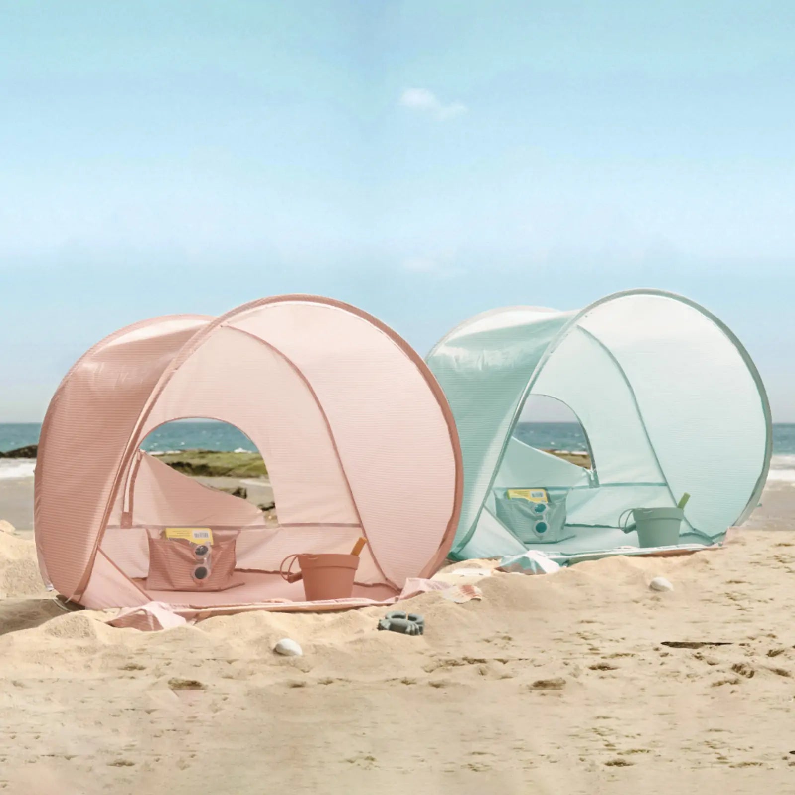 Two Tiny Land® Baby Beach Tents—Blush Pink and Light Blue—are set up on a sandy beach, each with toy buckets and shovels inside. These Tiny Land tents offer UV protection, with the ocean and blue sky in the background.