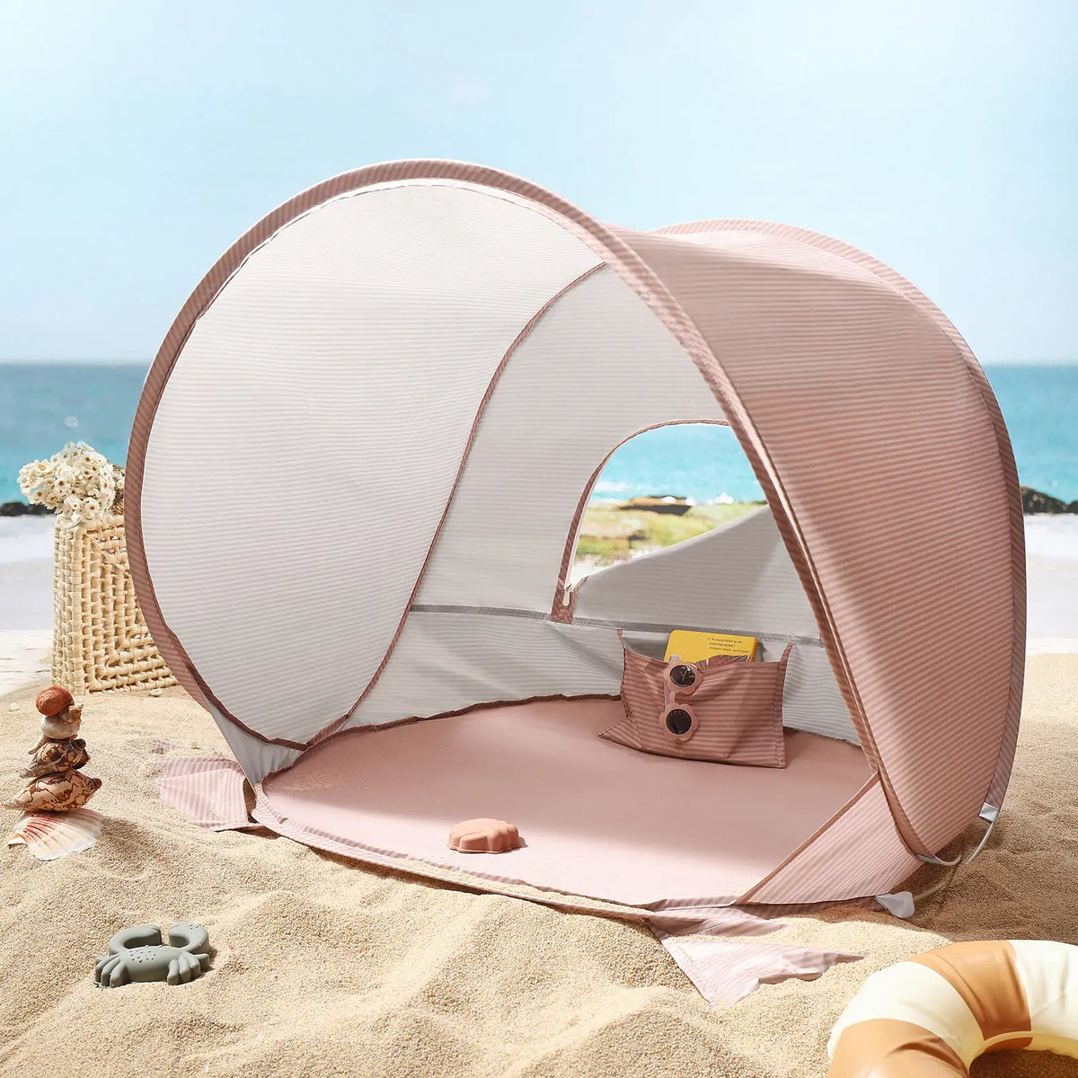 The Tiny Land® Baby Beach Tent in blush pink sits on the sand, offering UV protection with its open entrance facing the sea. Small toys and a wicker basket are nearby as waves roll in, inviting a cool breeze inside. Brand: Tiny Land.|Blush Pink