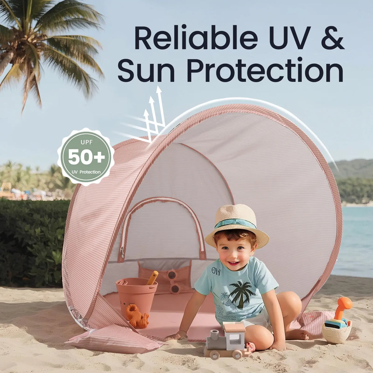A young child plays with toys on the sand inside the Tiny Land® Baby Beach Tent – Blush Pink, which offers UPF 50+ UV protection. The tent by Tiny Land is set against a scenic backdrop of palm trees and mountains.|Blush Pink