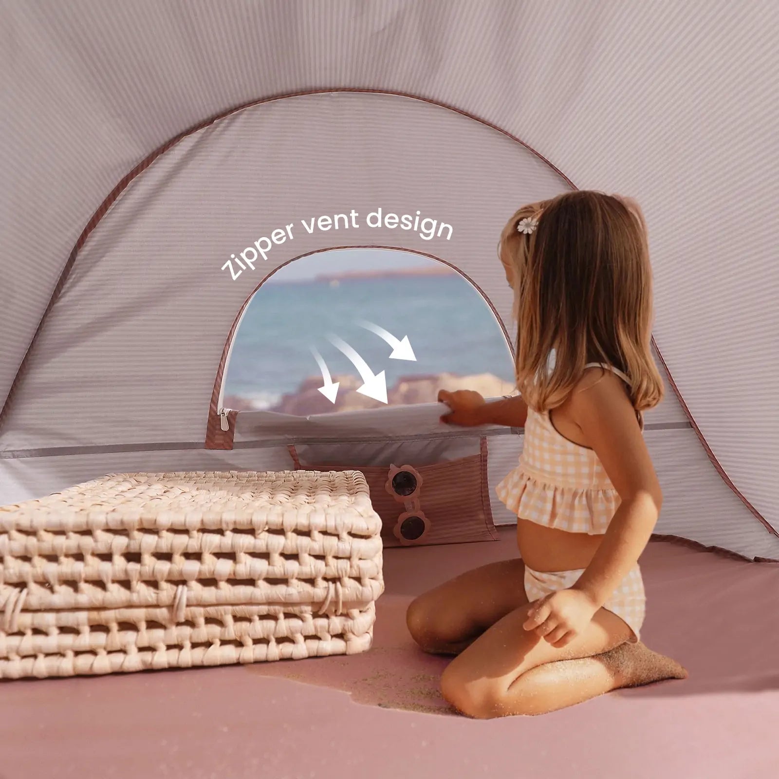 A young girl in a checked swimsuit enjoys the Tiny Land® Baby Beach Tent in blush pink on the sand, opening its zippered vent. Folded mats sit nearby, with the sea visible through the window. “Zipper vent design” is displayed in white text.