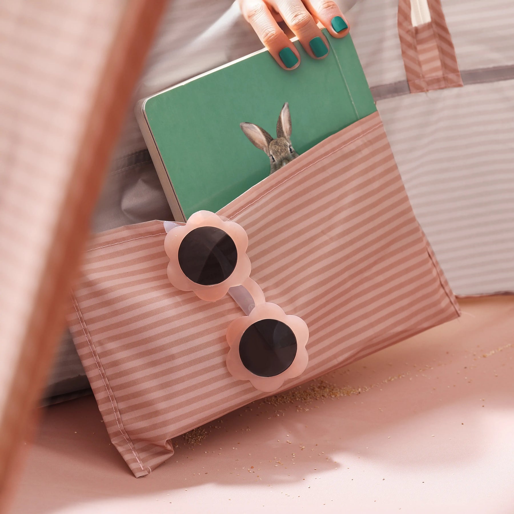 A hand packs a green notebook with a rabbit cover into a pink-and-white pouch alongside flower-shaped sunglasses—ideal for unwinding by the Tiny Land® Baby Beach Tent – Blush Pink Play Tent. Sand is sprinkled across the light pink surface.