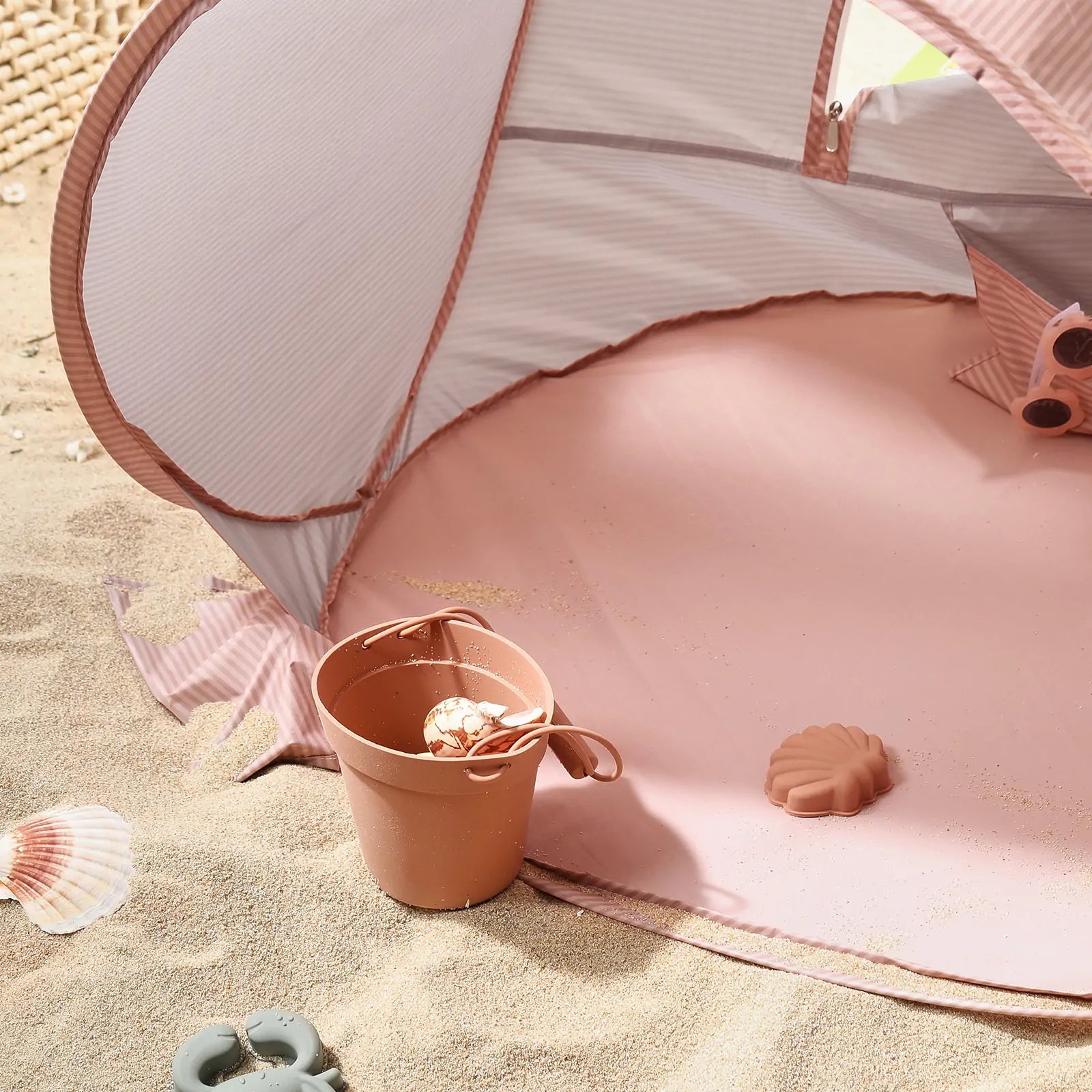 The Tiny Land® Baby Beach Tent in blush pink is set up on the sand, with a brown bucket holding a seashell and toy mold nearby. Scattered seashells complete this summery outdoor scene by Tiny Land.