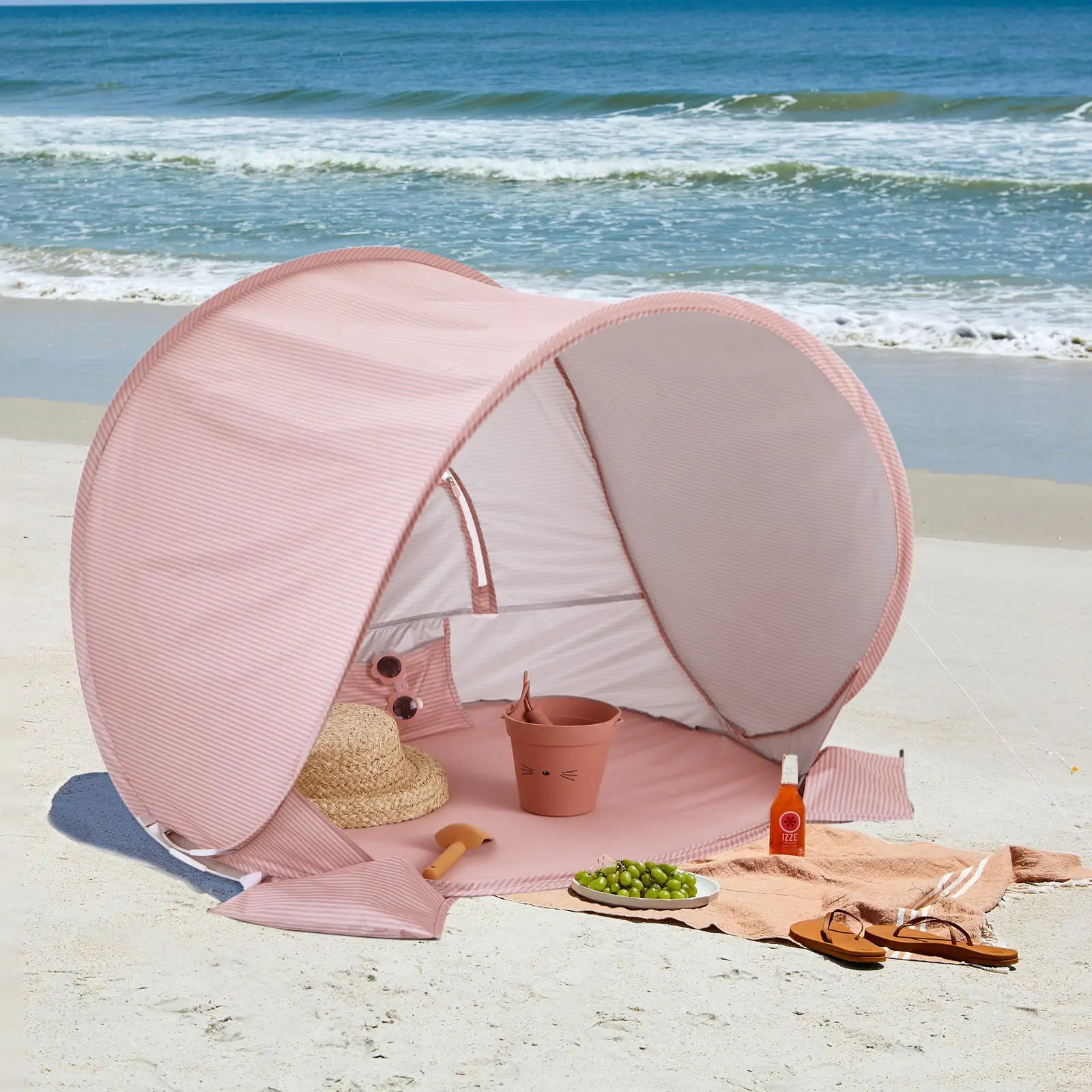The Tiny Land® Baby Beach Tent in blush pink sits on the sand near the ocean. Inside are a straw hat, pail, sunglasses, and grapes; outside, sandals, sunscreen, and a towel await under rolling waves and a blue sky.
