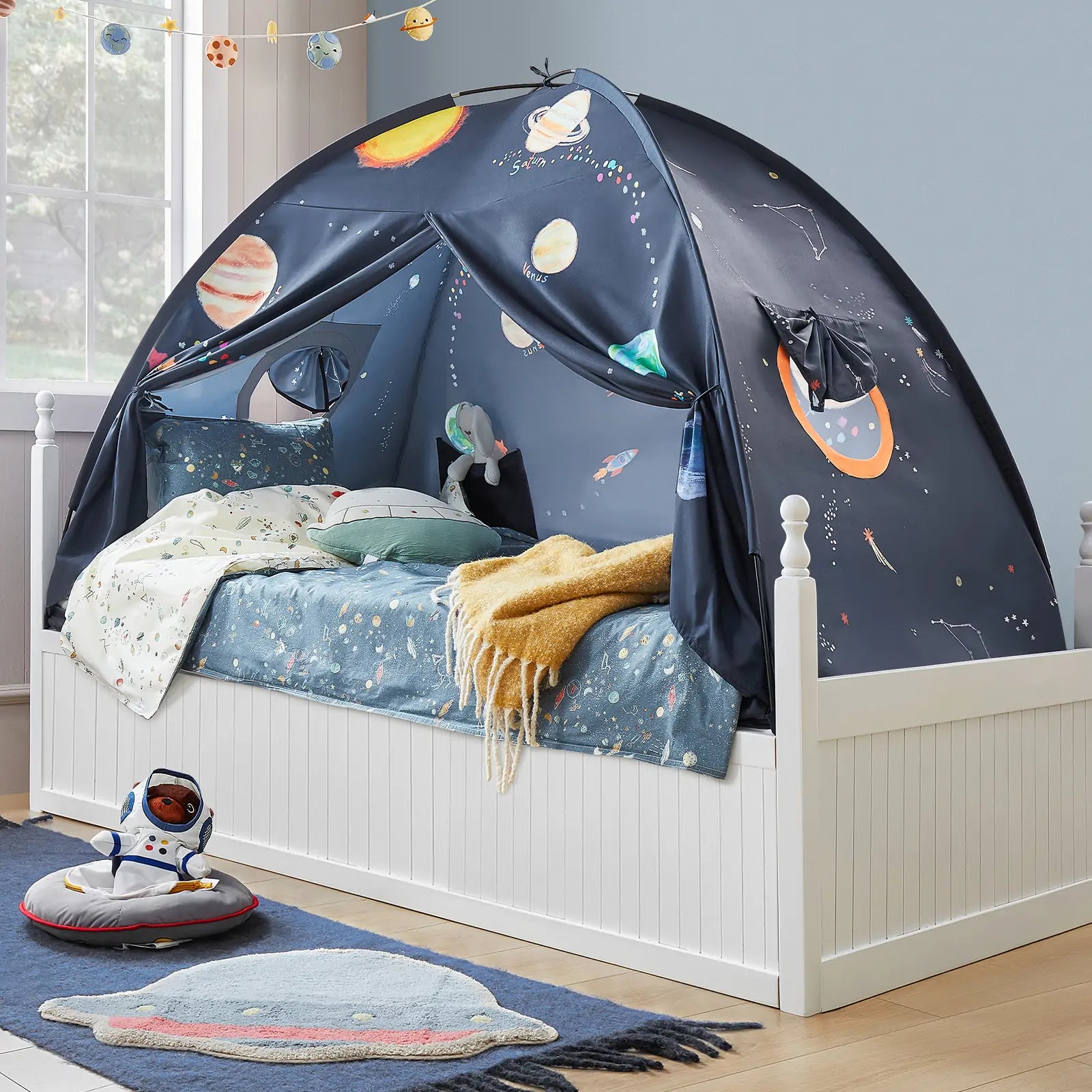 The Tiny Land® SpaceNest Bed Tent by Tiny Land INC, featuring planets and space designs, is set up in a bright room with light blue space-themed bedding and a plush astronaut on a planet-shaped rug nearby.