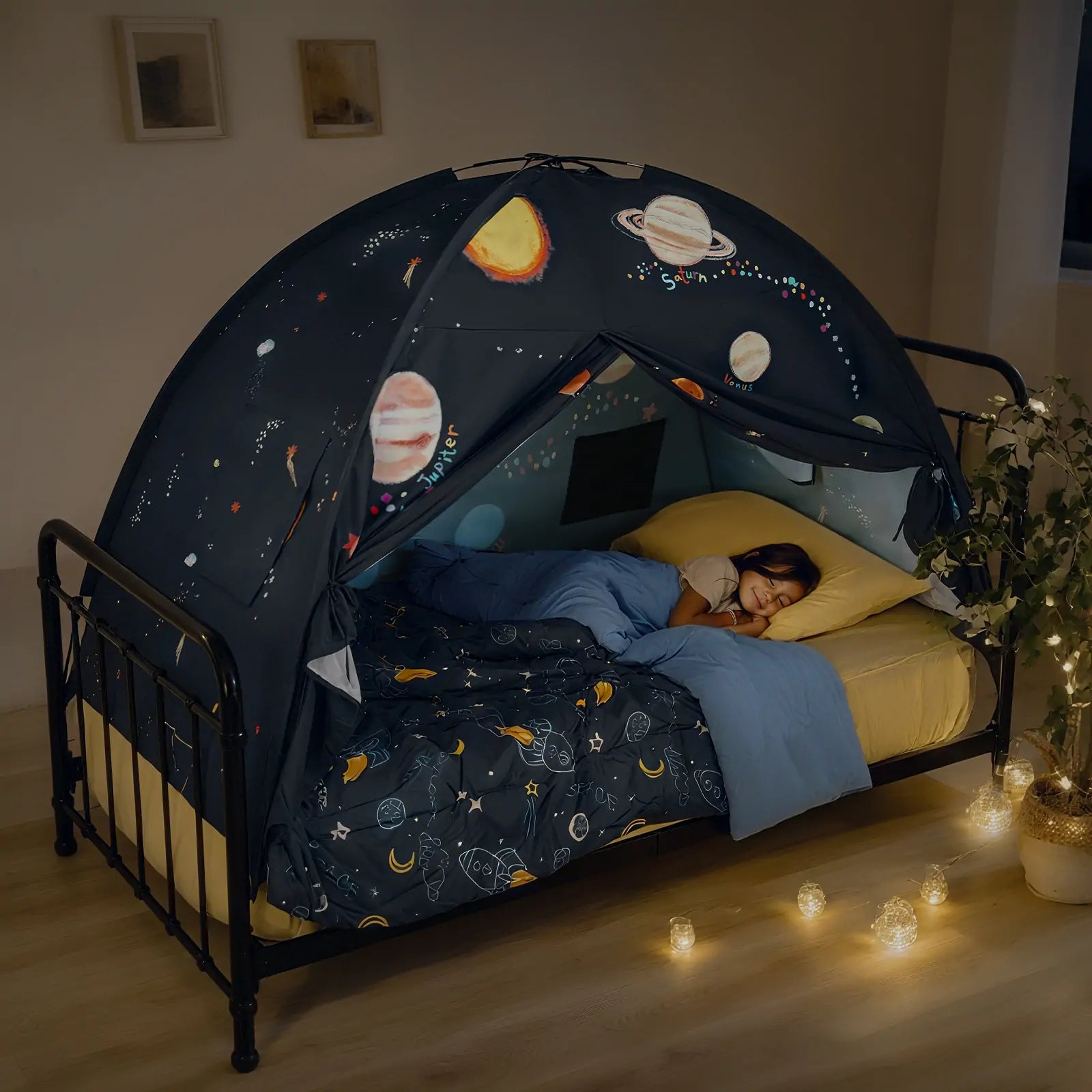 A child sleeps in a Tiny Land® SpaceNest Bed Tent by Tiny Land INC, enjoying an indoor adventure under planets and stars. Soft string lights illuminate the dim room and a potted plant adds a touch of life nearby.