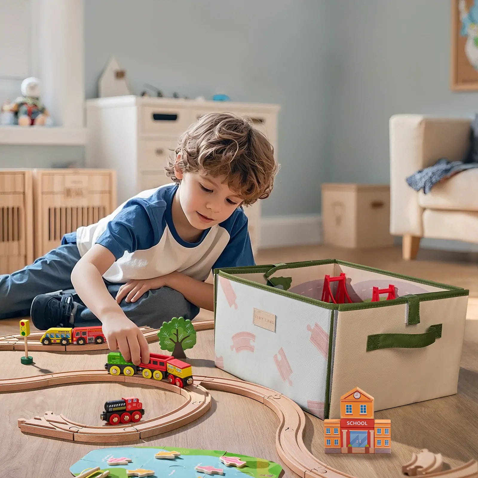 A young boy with curly hair plays on the floor with the Tiny Land® 110-Piece Wooden Train Set and its folding storage box, beside a city play mat in a bright playroom. Wooden crates and a small couch are seen in the background.